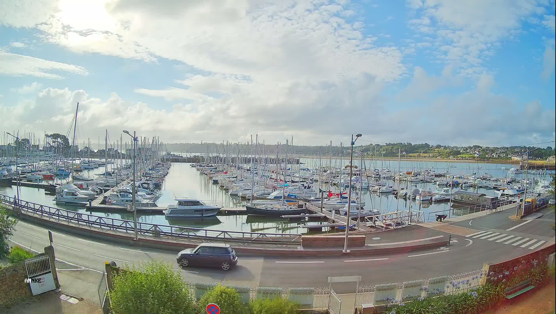 A marina filled with numerous sailboats and motorboats is visible under a partly cloudy sky, with a small car driving on a road next to it.