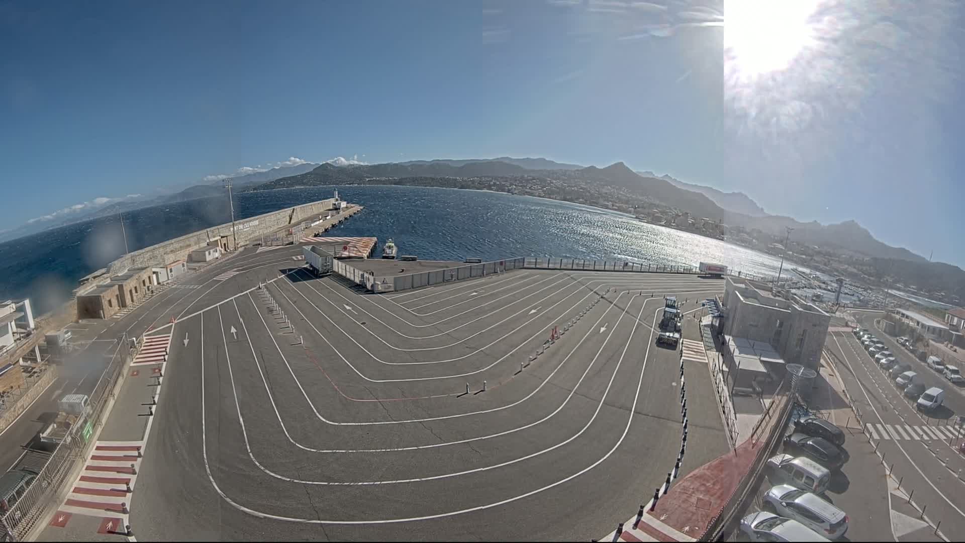 A large, mostly empty vehicle staging area with marked lanes next to a body of water under a sunny sky, with mountains visible in the distance.