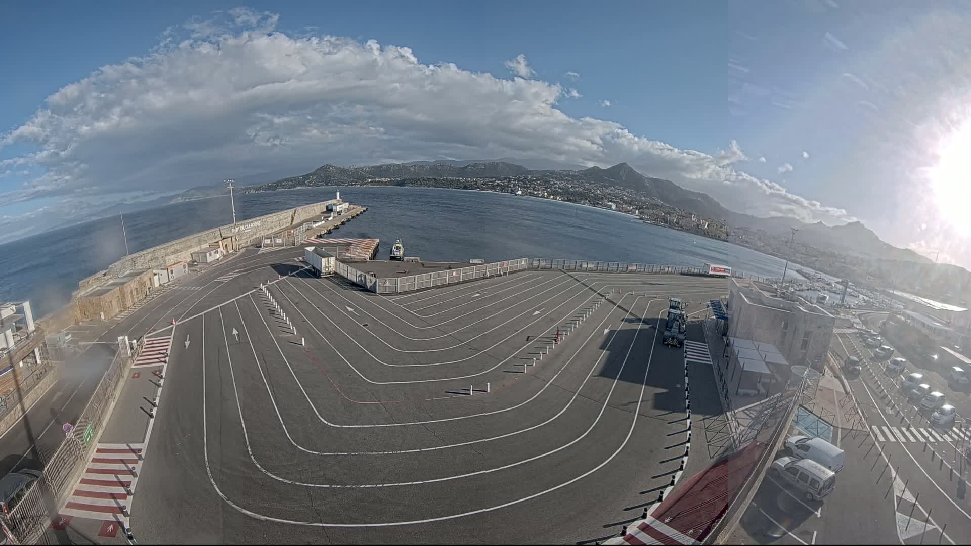 An empty ferry terminal with winding vehicle lanes overlooks a calm sea under a partly cloudy sky, with mountains visible in the distance.