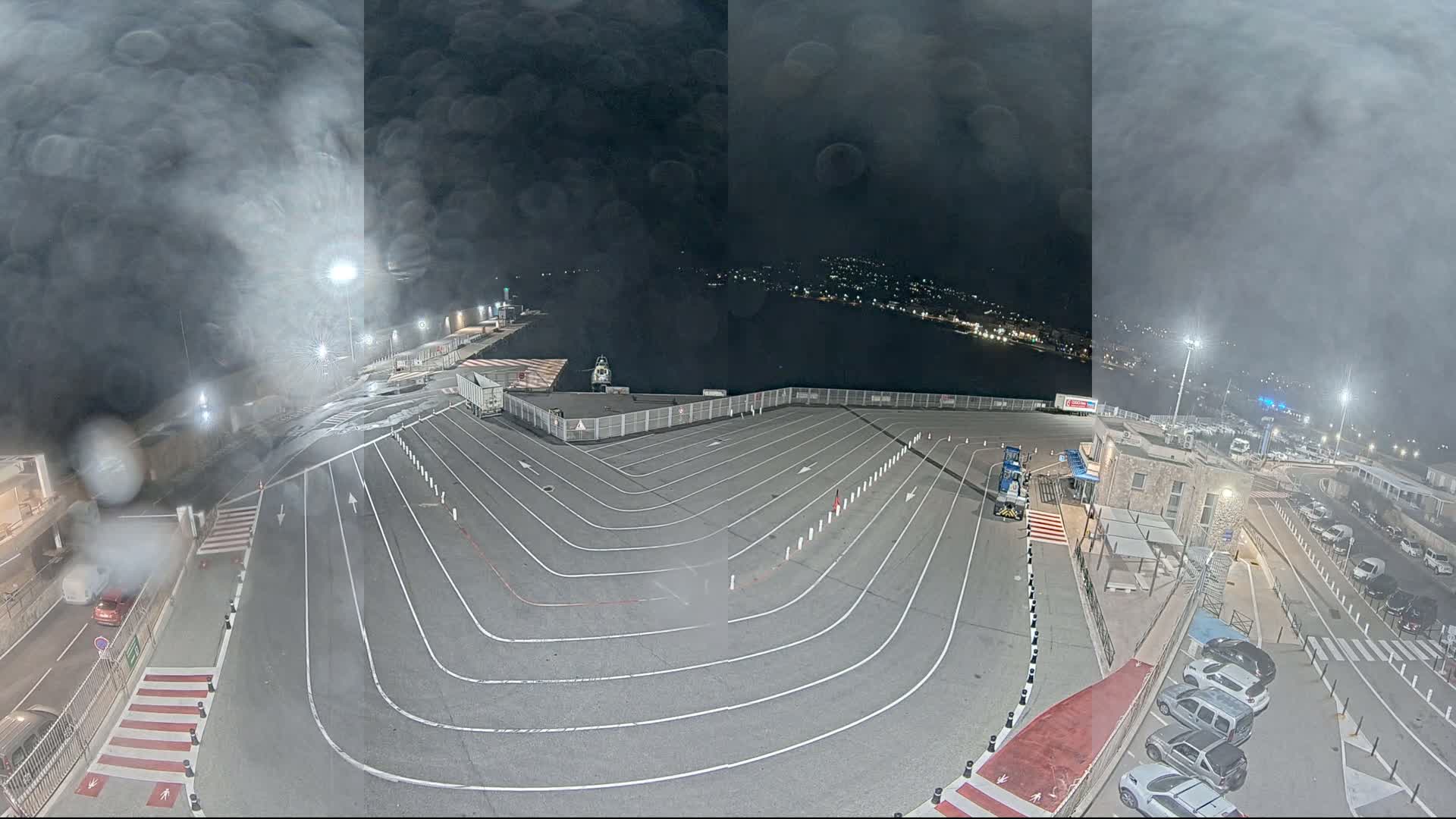 A nighttime aerial view of a port area with a large, marked parking lot, several vehicles, and a body of water visible in the background under a hazy, cloudy sky.