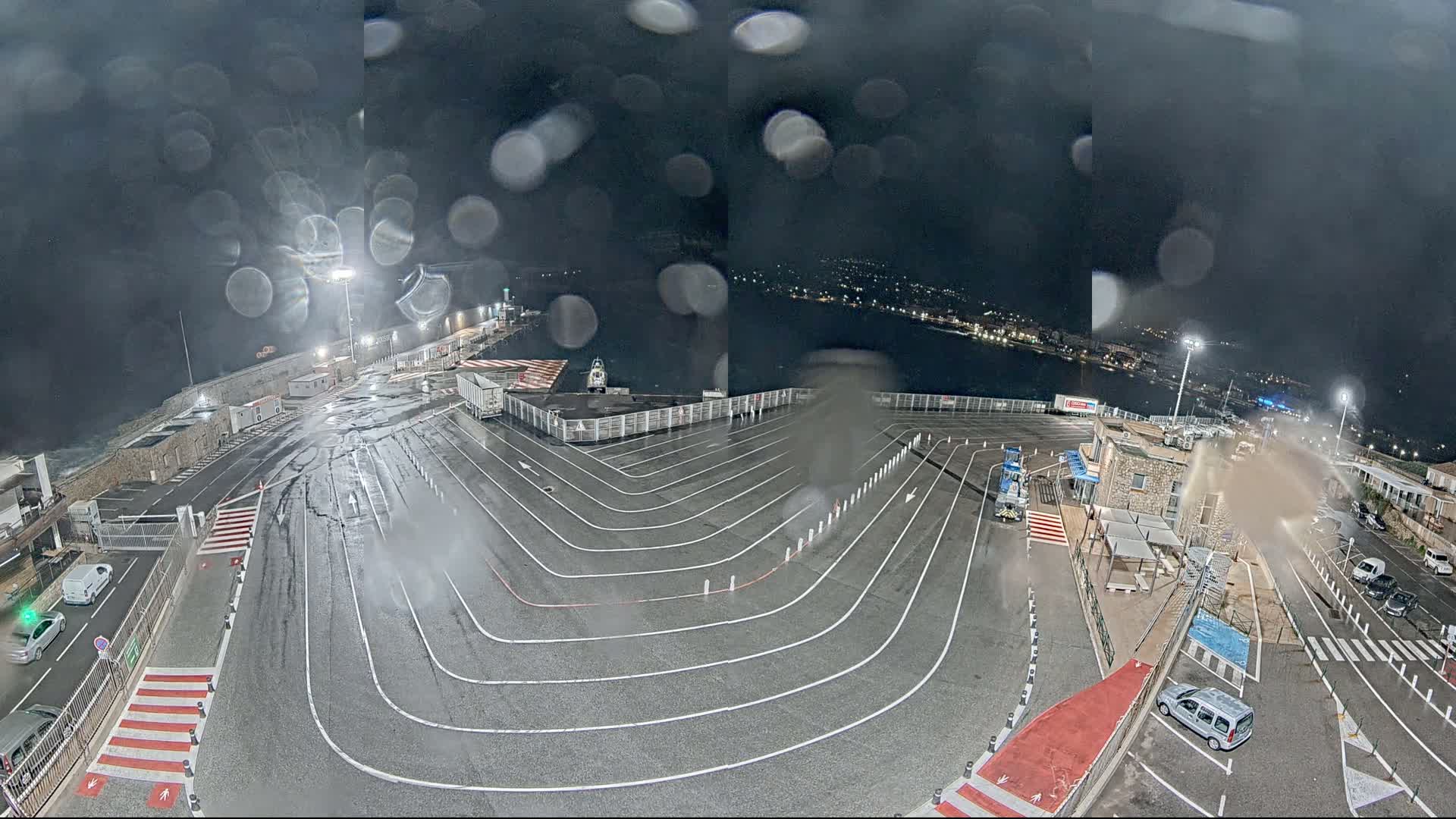 A rain-slicked ferry terminal at night, with a curving vehicle queuing area, is seen through rain-covered glass.