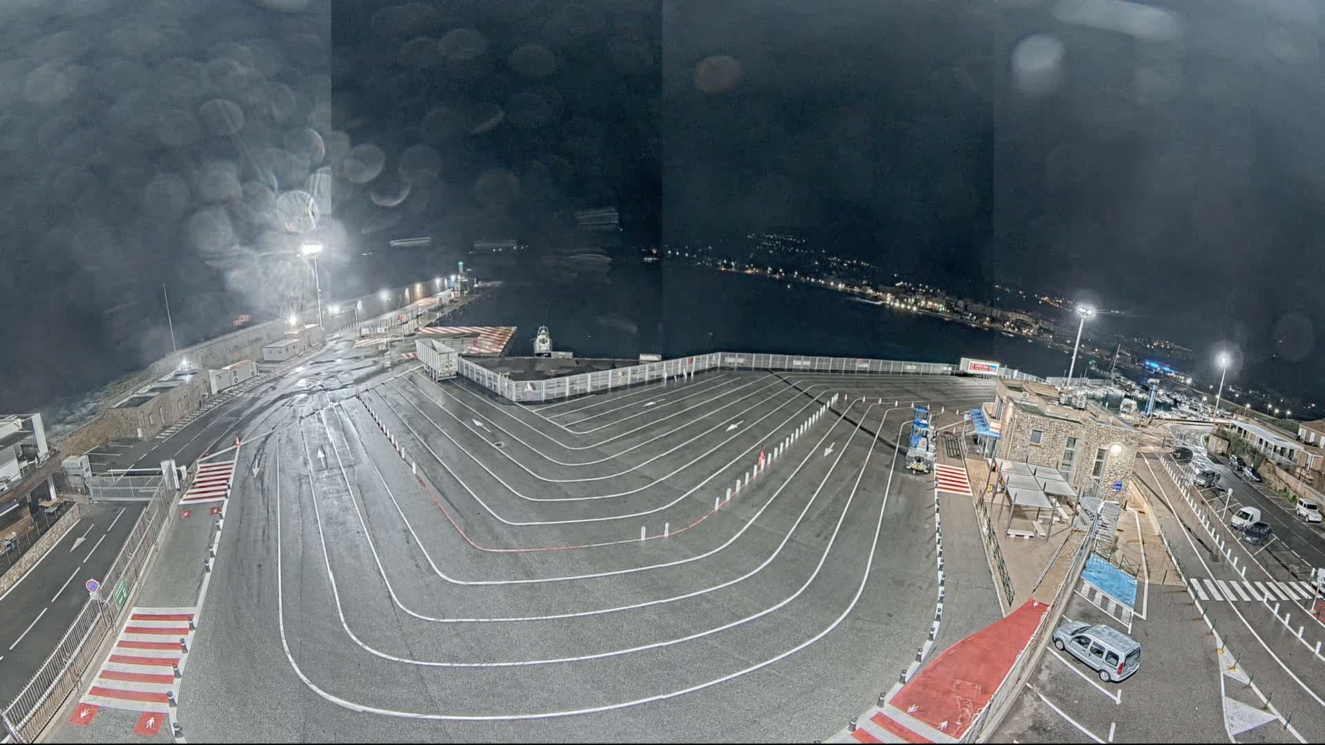 A wet, nighttime view of a ferry terminal with a large, marked vehicle queueing area overlooking a dark body of water.