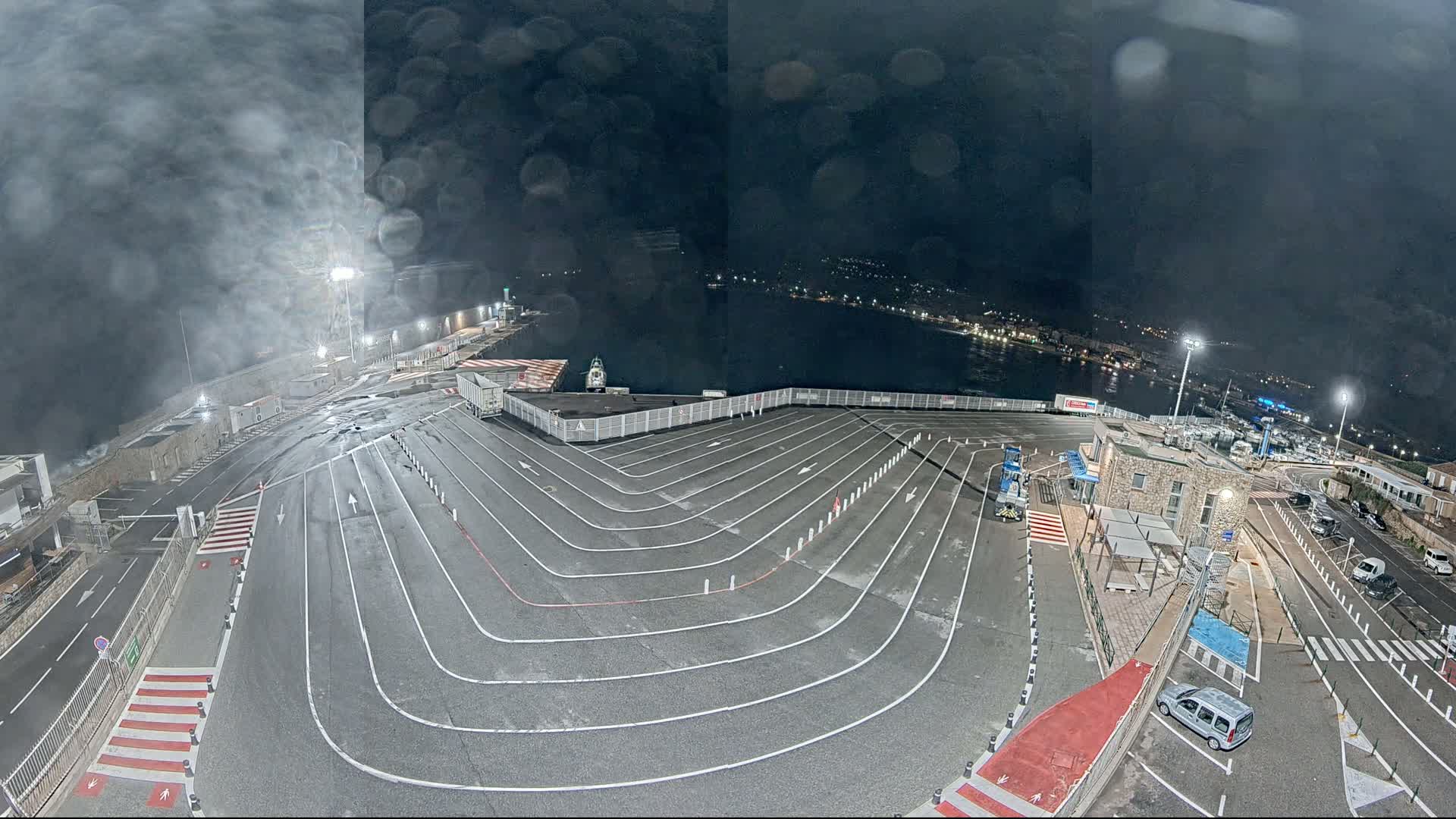 An empty, winding ferry terminal at night under a hazy, dark sky, with a few vehicles visible in the surrounding parking area and a body of water in the background.