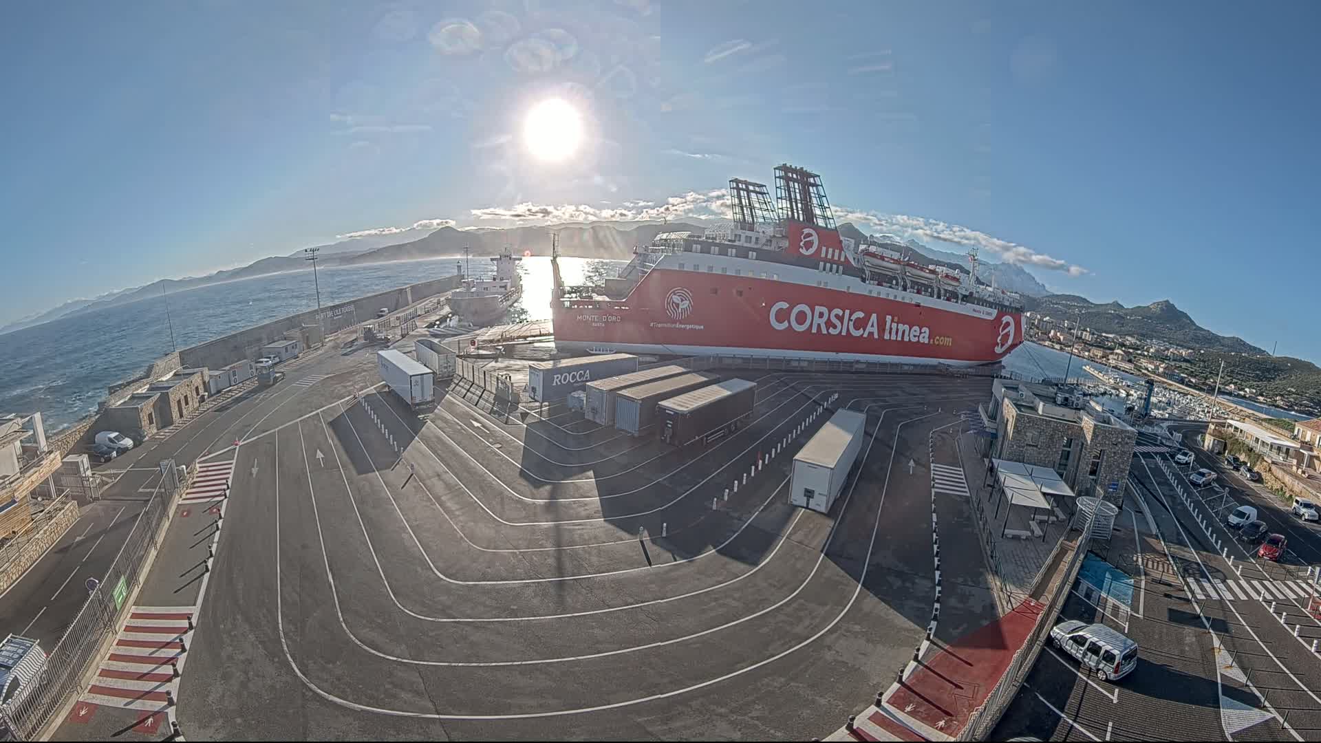 A large red ferry is docked at a port on a sunny day with calm waters and mountains in the background.