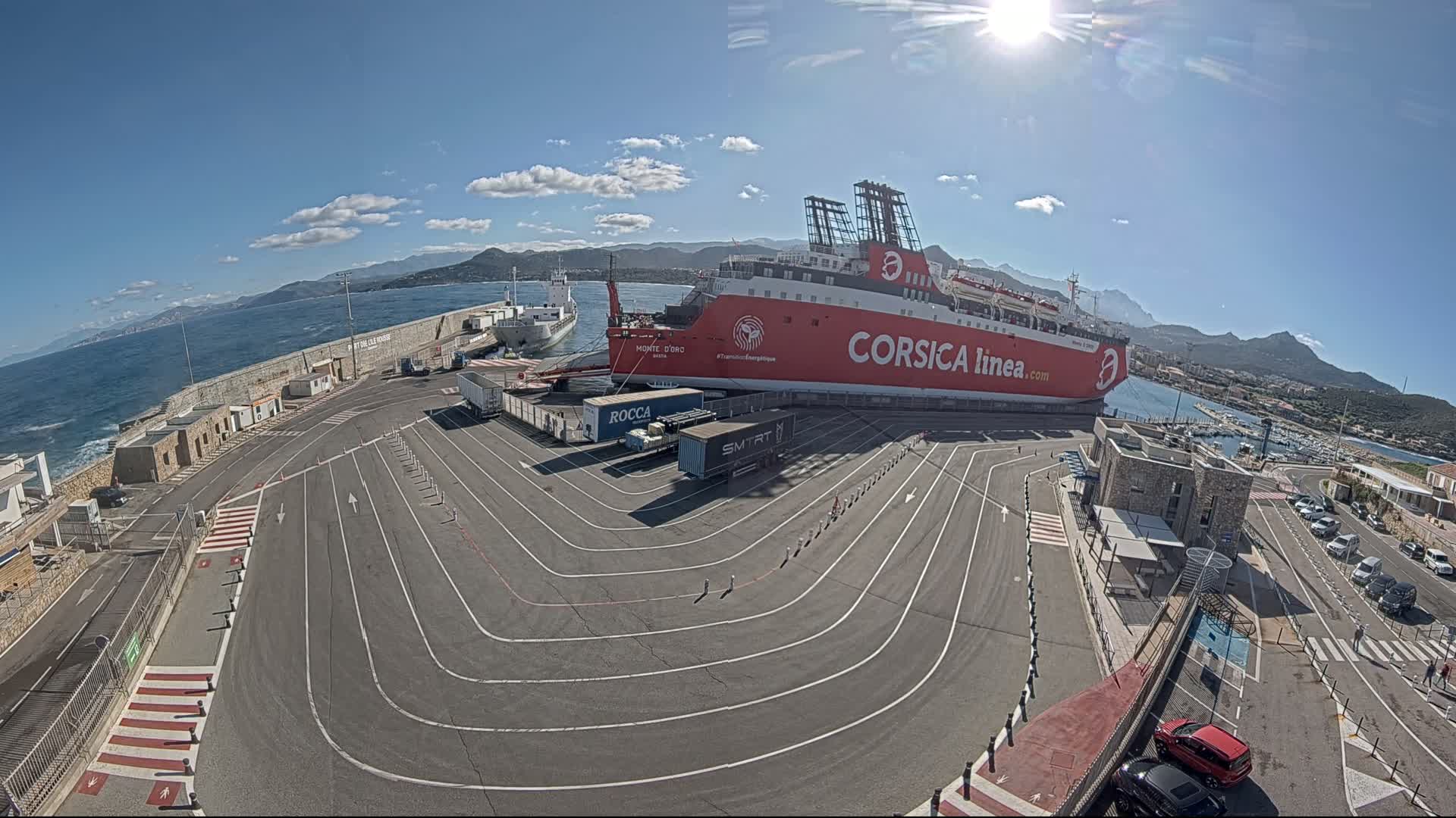 A large red ferry is docked at a port on a sunny day, with a smaller cargo ship nearby and several trucks and cars in the parking lot.