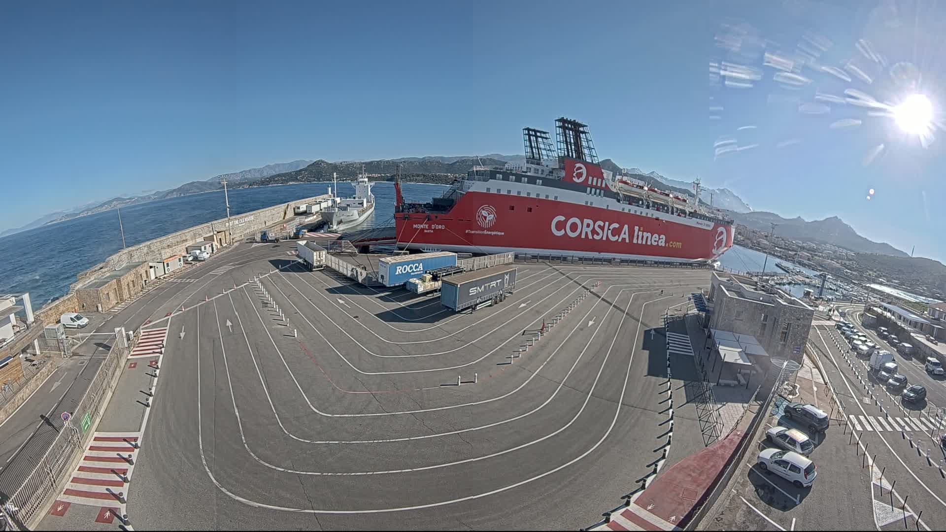 A large red ferry is docked at a port on a sunny day, with several trucks and cars parked nearby, against a backdrop of mountains and calm blue water.