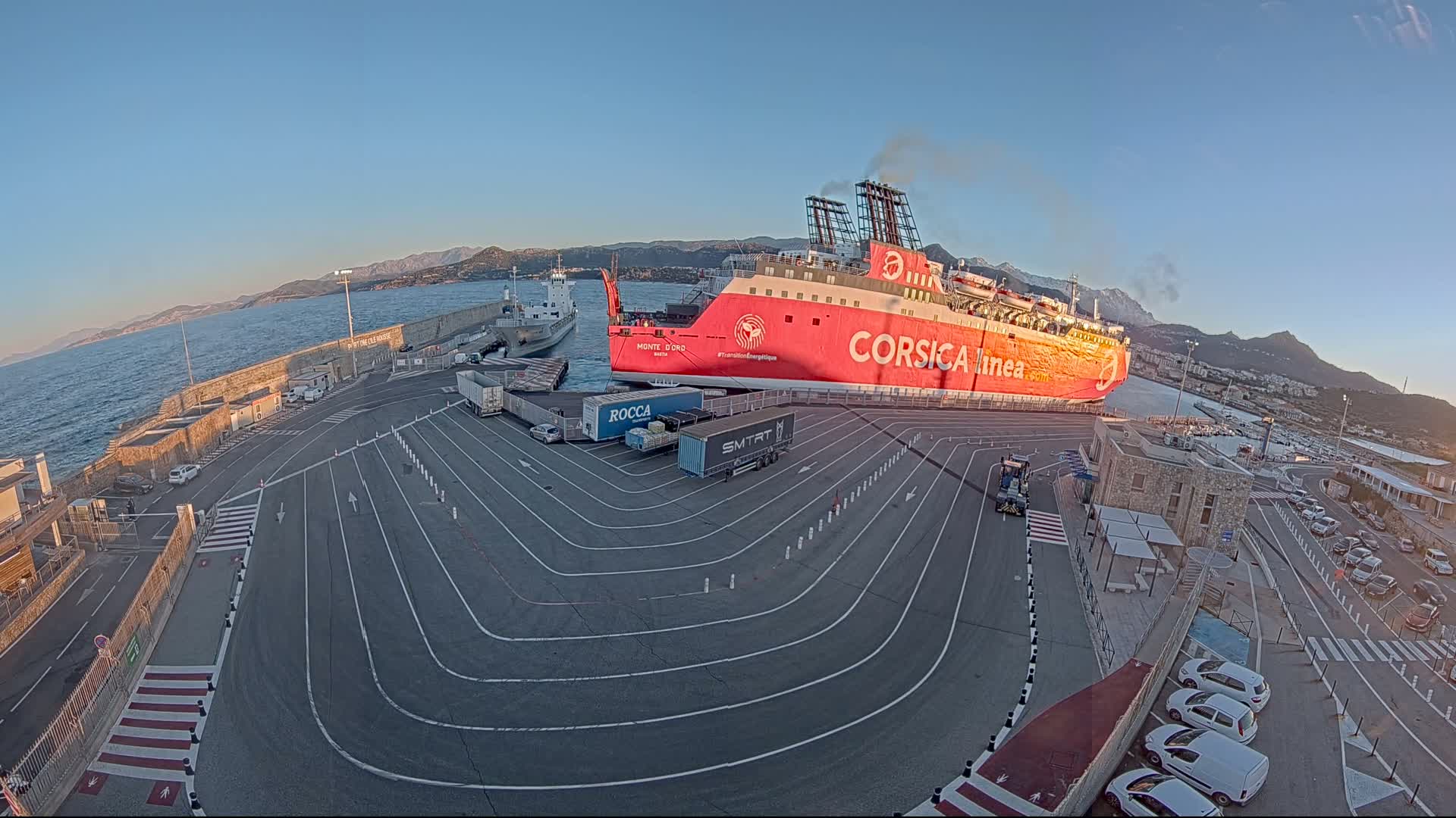 A large red ferry is docked at a port, under a clear, bright sky, with several cars and trucks parked nearby.