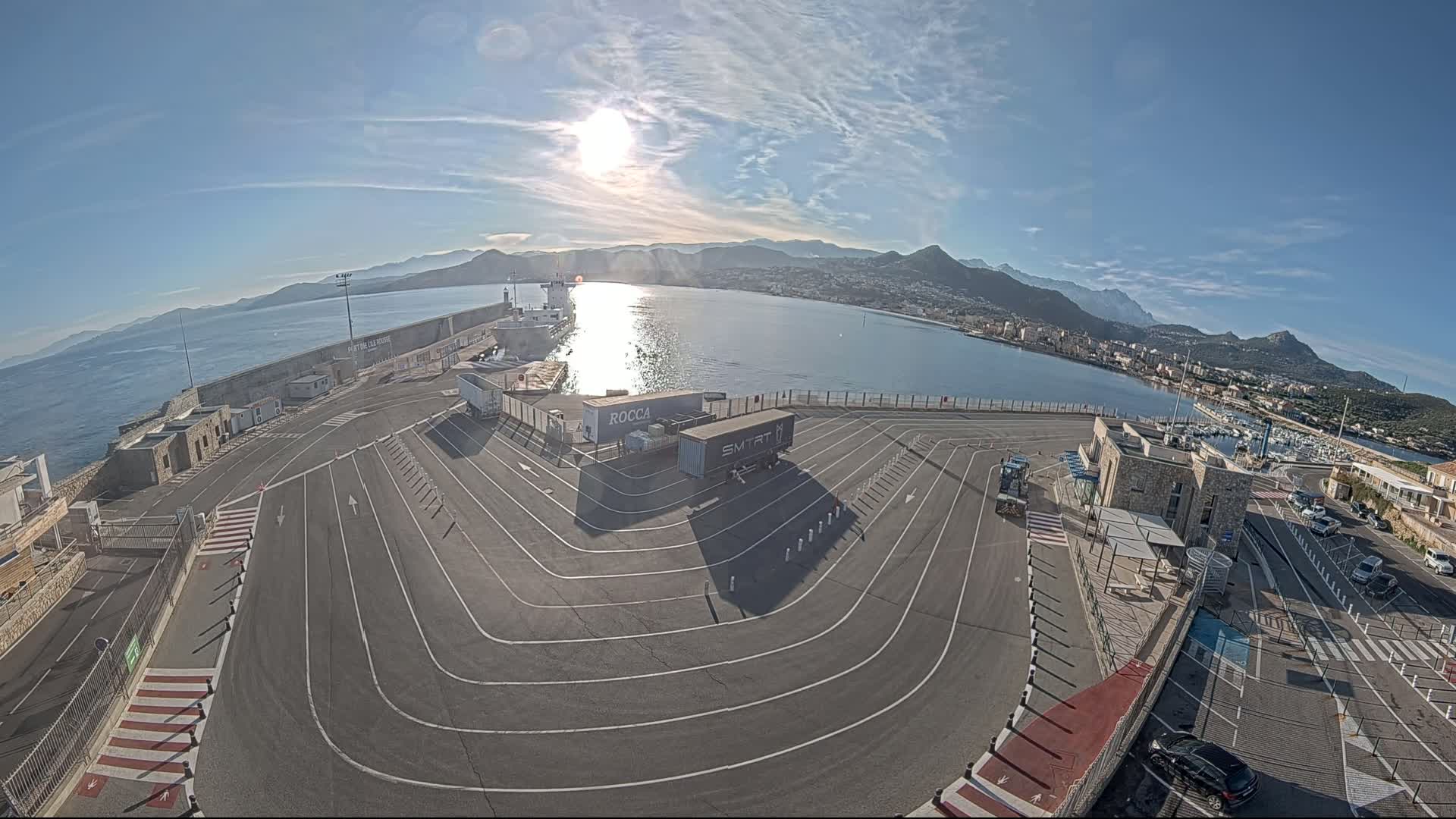 A sunny day at a harbor shows a large paved area with marked lanes, several trucks, and a small building, all overlooking calm water with mountains in the distance.