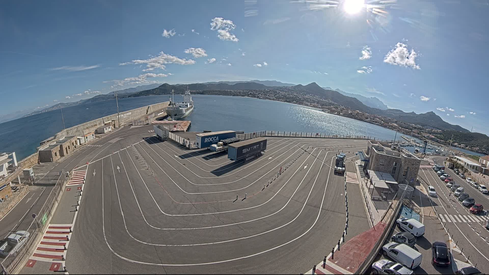 A sunny day at a harbor shows a large ship docked, several trucks in a parking lot with marked lanes, and cars parked nearby, all against a backdrop of mountains and calm water.
