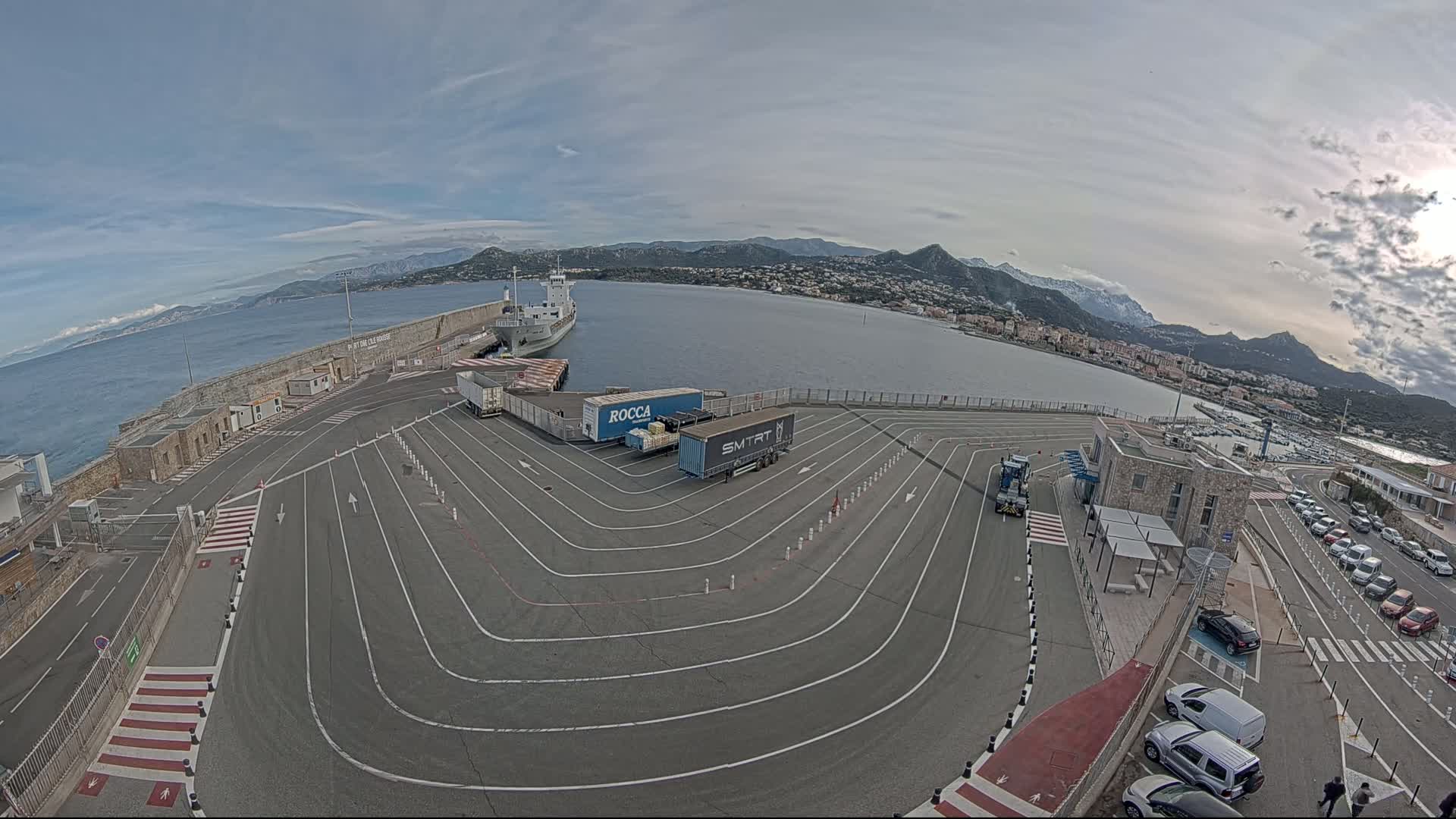 A mostly clear day at a port with a cargo ship docked, several trucks in a paved turning area, and cars parked in a lot overlooking a calm body of water and mountains in the distance.