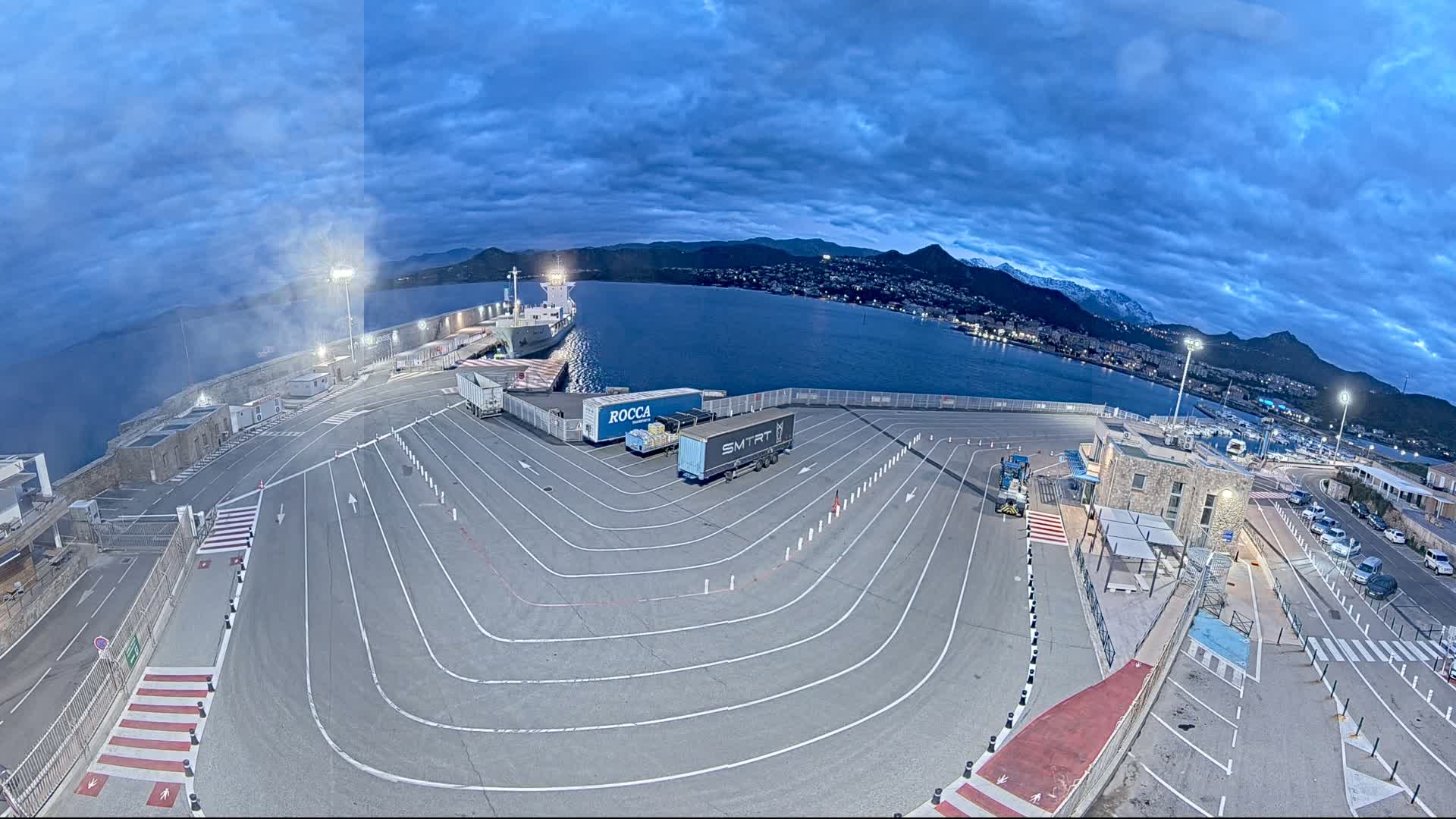 An empty port at dusk, under a cloudy sky, with a large ship docked and several trucks parked on the tarmac.