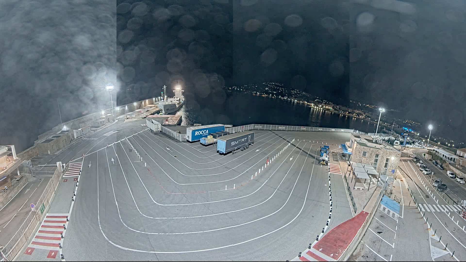 A nighttime aerial view of a port shows a cargo ship and several trucks in a parking lot with marked lanes, under a cloudy sky.