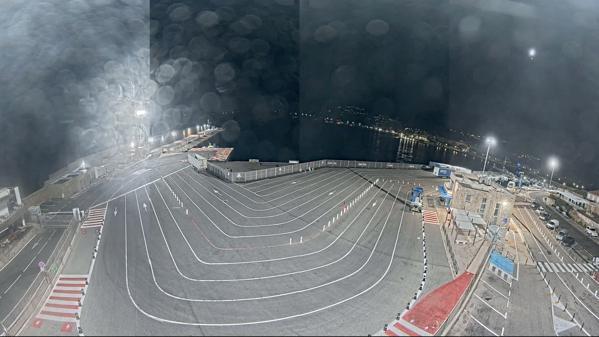 A nighttime view of a ferry terminal with raindrops on the camera lens, showing a large, empty vehicle staging area with marked lanes next to a body of water.