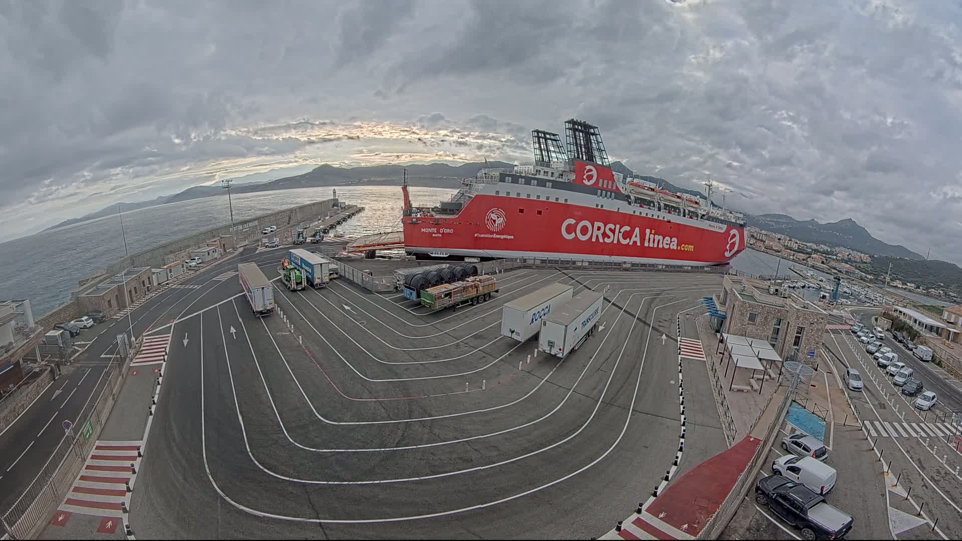 A large red ferry is docked at a port under a cloudy sky, with several trucks and cars in the surrounding parking lot.