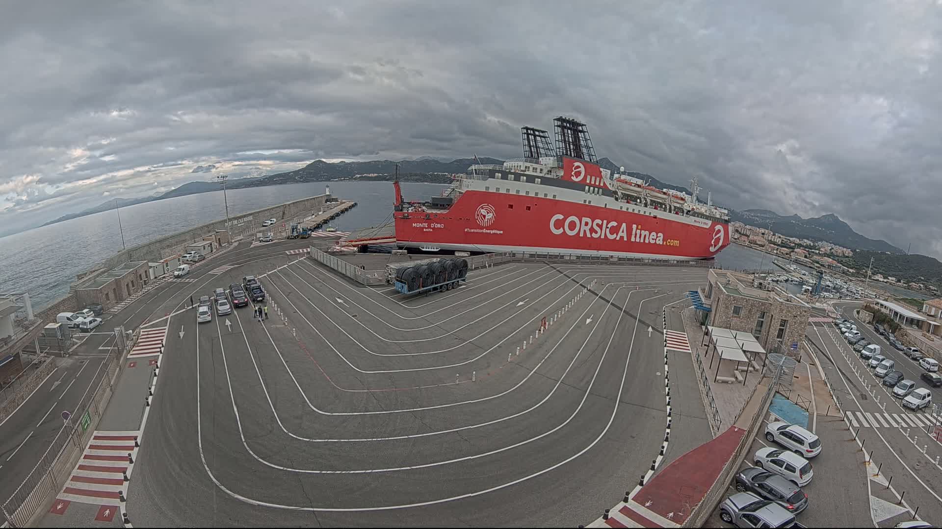 A large red ferry is docked at a port on an overcast day, with several cars parked in the surrounding area.