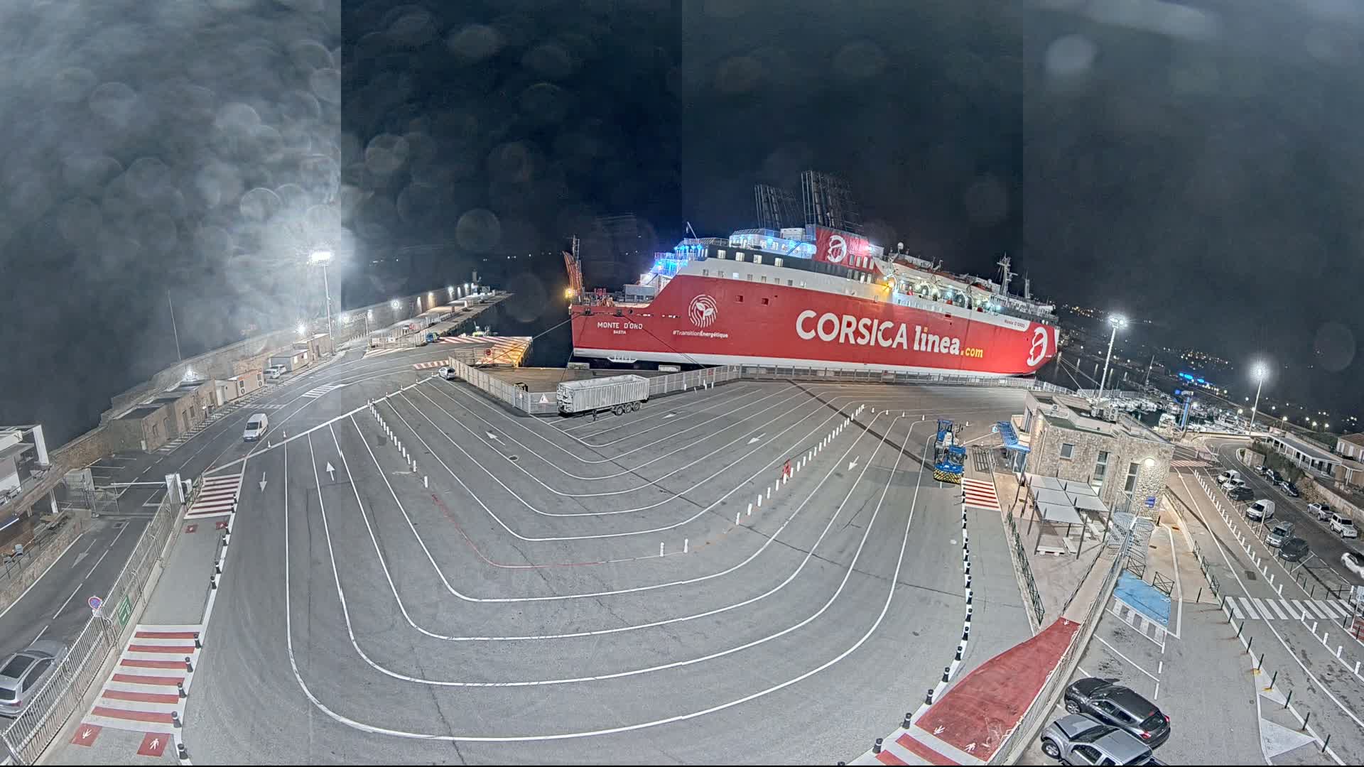 A large red ferry is docked at night in a port with a dark, overcast sky, and the port is mostly empty aside from a few cars.