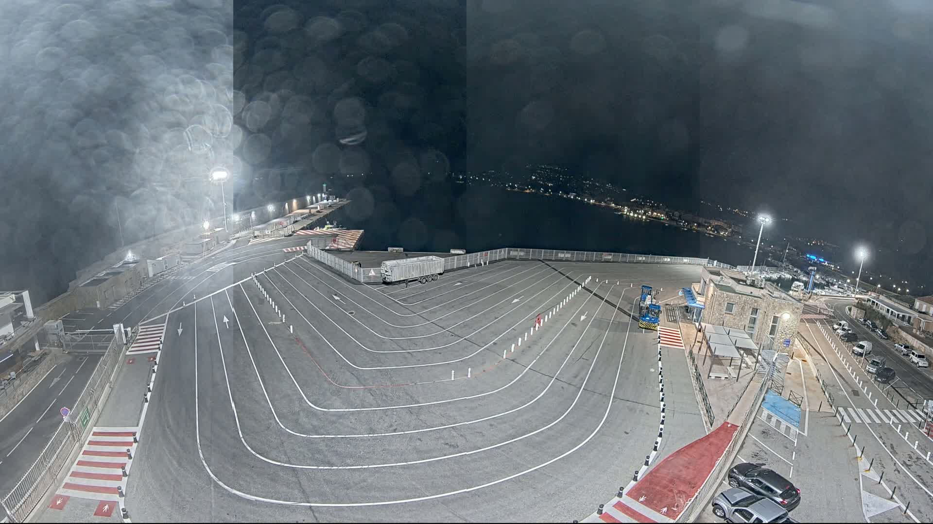 A nighttime, rain-streaked view of a large, empty parking lot with marked lanes at a waterfront location, overlooking a dark body of water and a town in the distance.