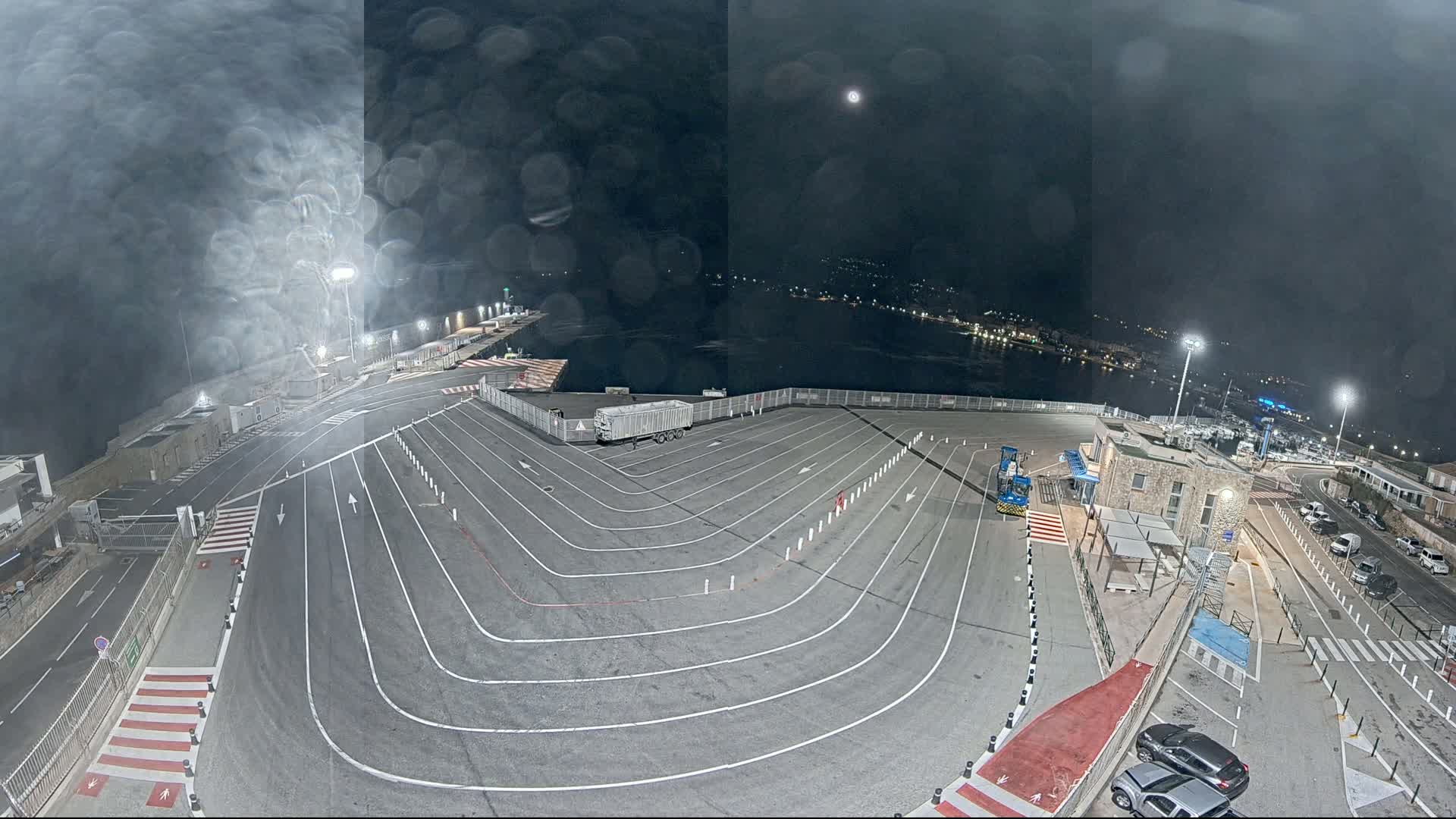 An empty, lined parking lot at night overlooks a dark body of water under a slightly cloudy sky, with a few cars parked nearby.
