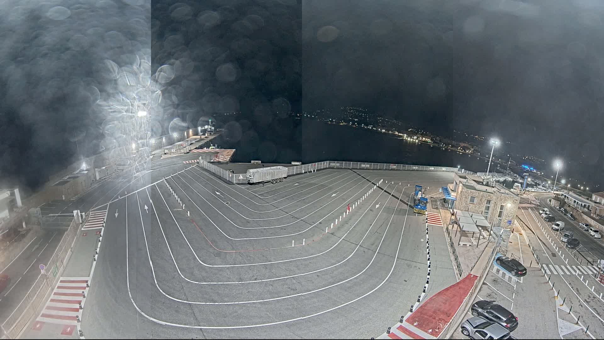 A nighttime view of a ferry terminal with a large, empty vehicle holding area marked with painted lines, under a dark, rainy sky.