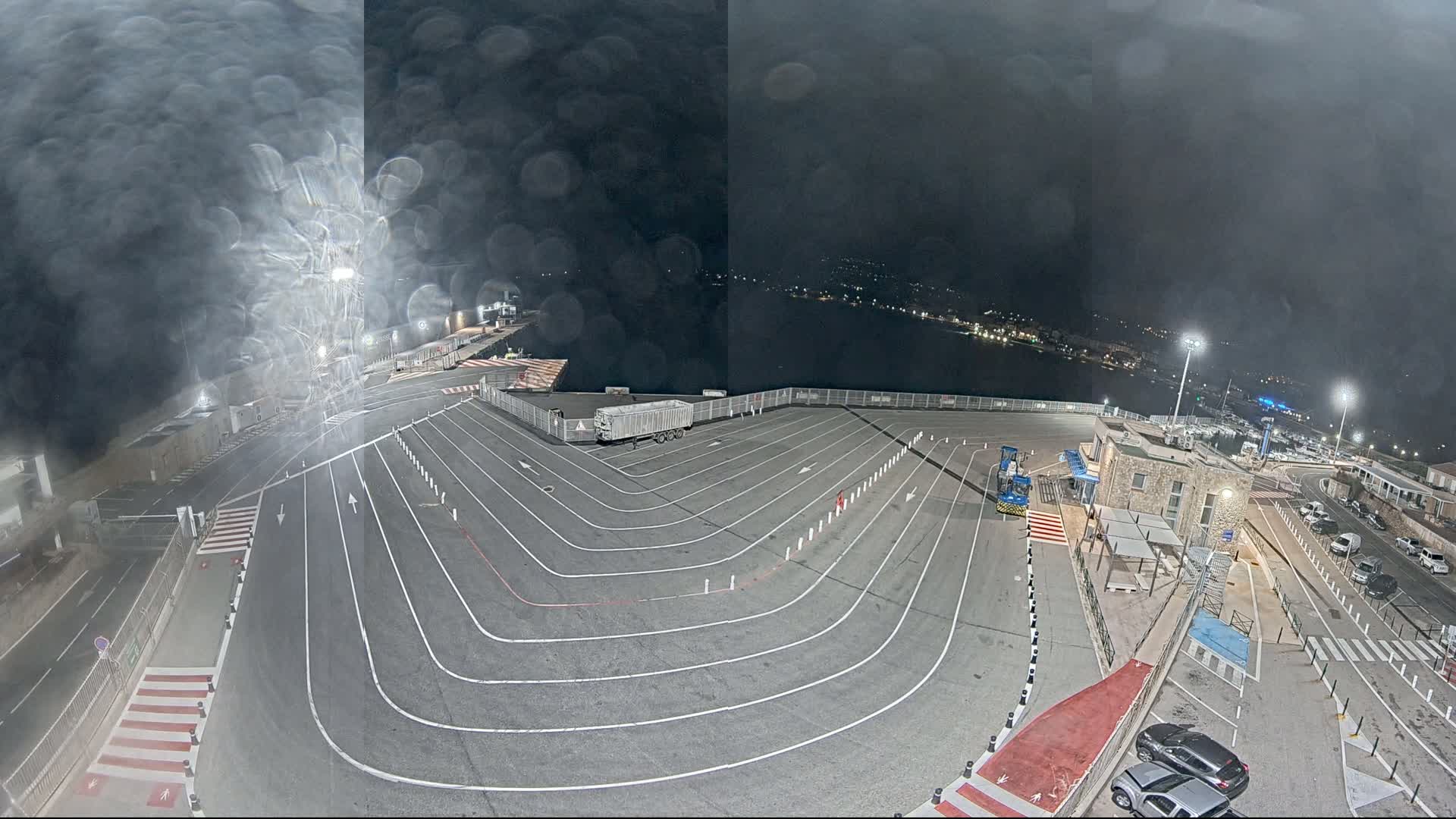 A nighttime aerial view of a ferry terminal's vehicle staging area shows a large, empty lot with painted lines and a few parked cars, under a dark, rain-streaked sky.