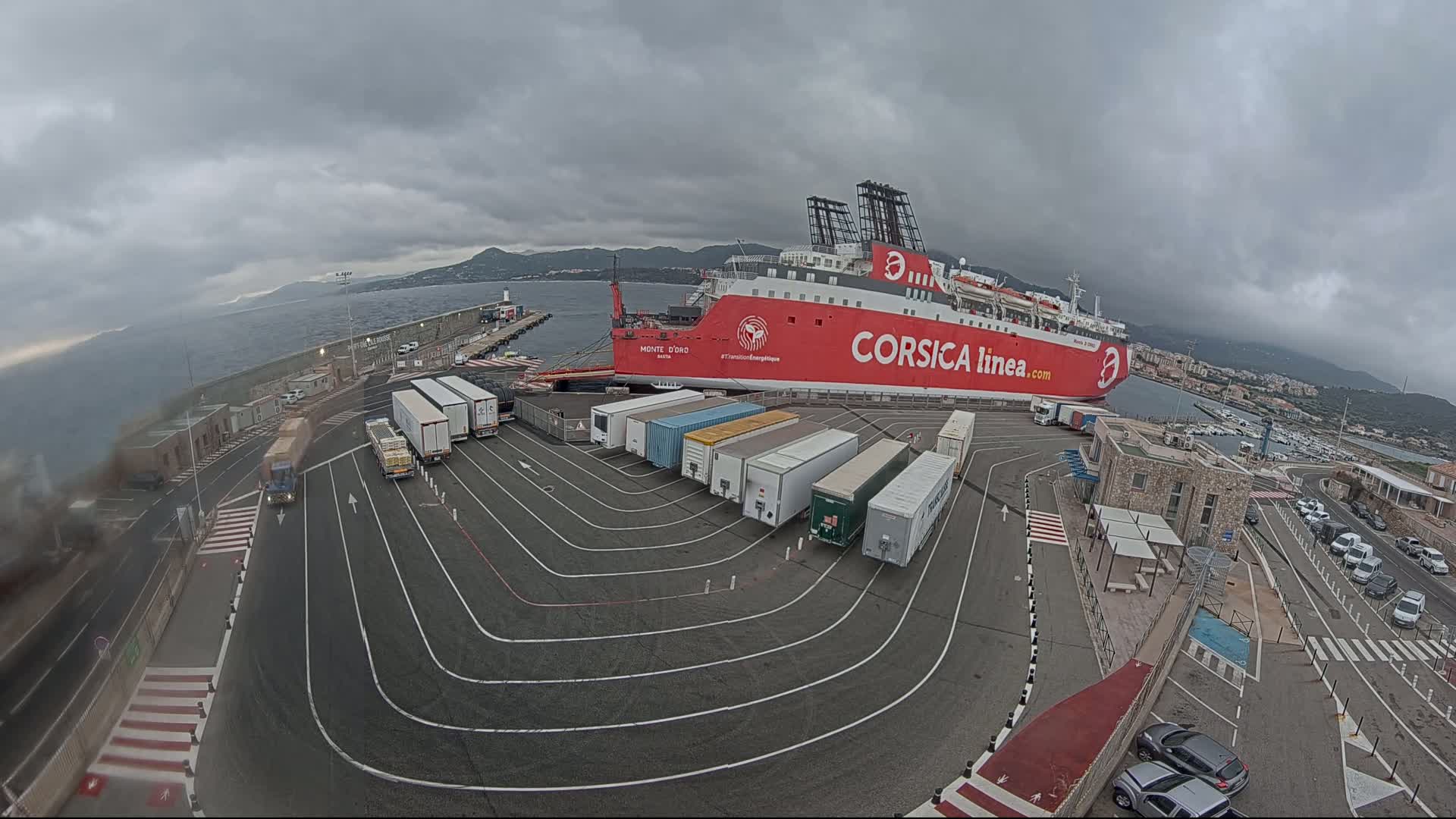 A large red ferry is docked at a port under an overcast sky, with numerous trucks and trailers lined up nearby.