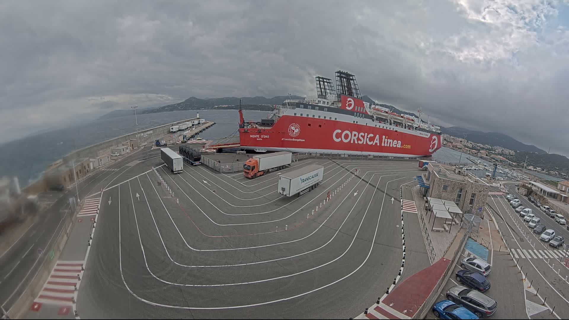 A large red ferry is docked at a port under an overcast sky, with several trucks and cars parked nearby.