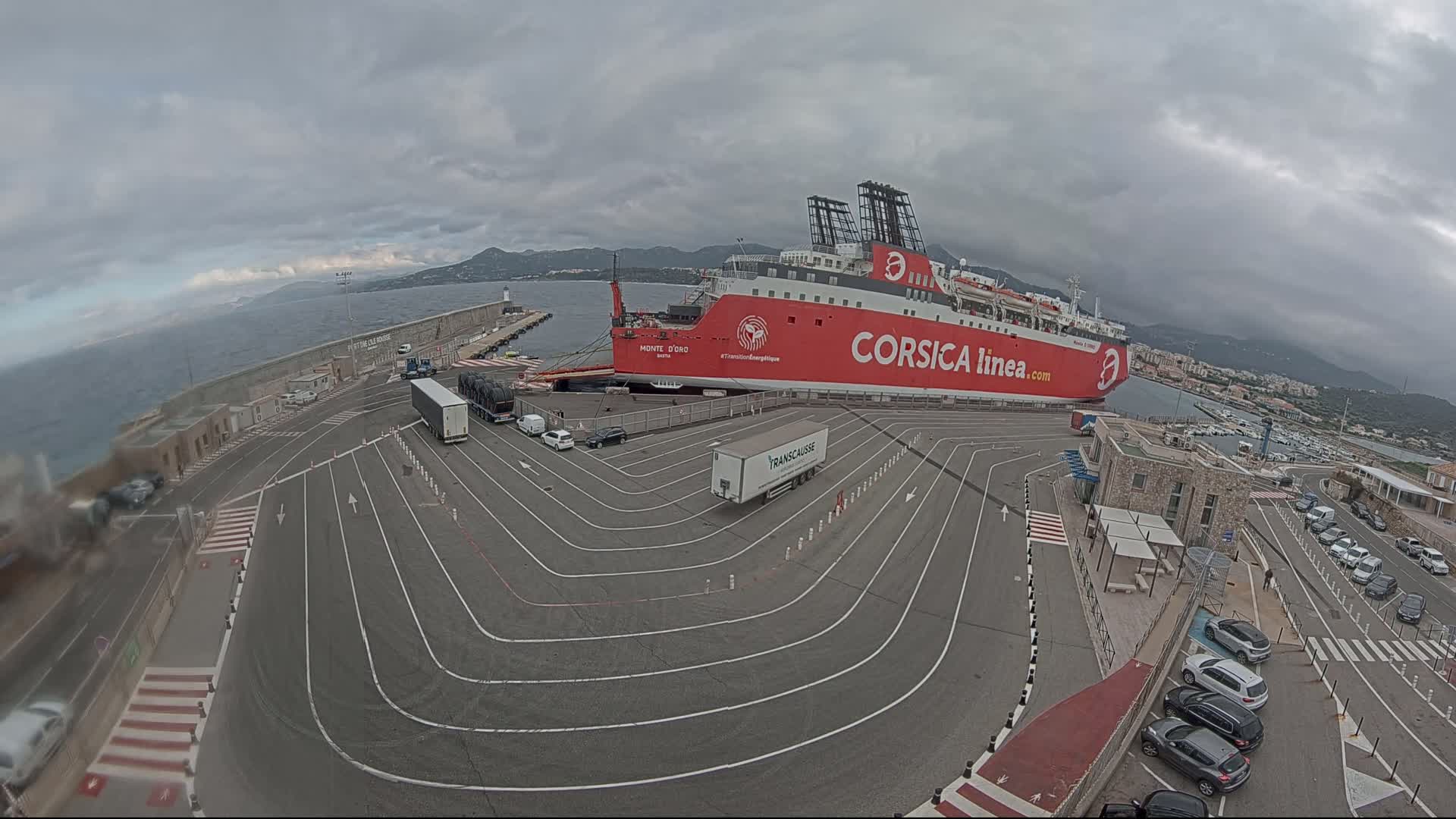 A large red ferry is docked at a port under an overcast sky, with several vehicles parked nearby in designated lanes.