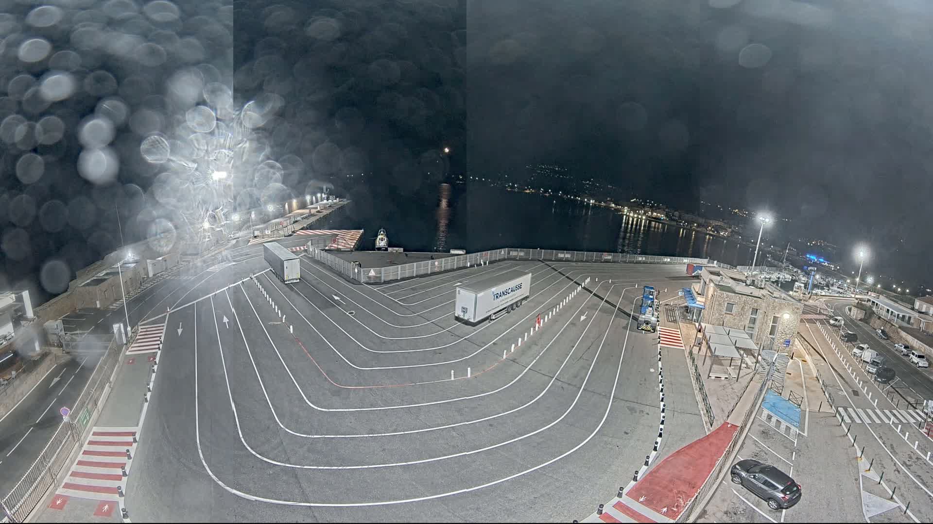 A nighttime view of a port area with marked lanes, a few vehicles, and a body of water visible in the background under rainy conditions.