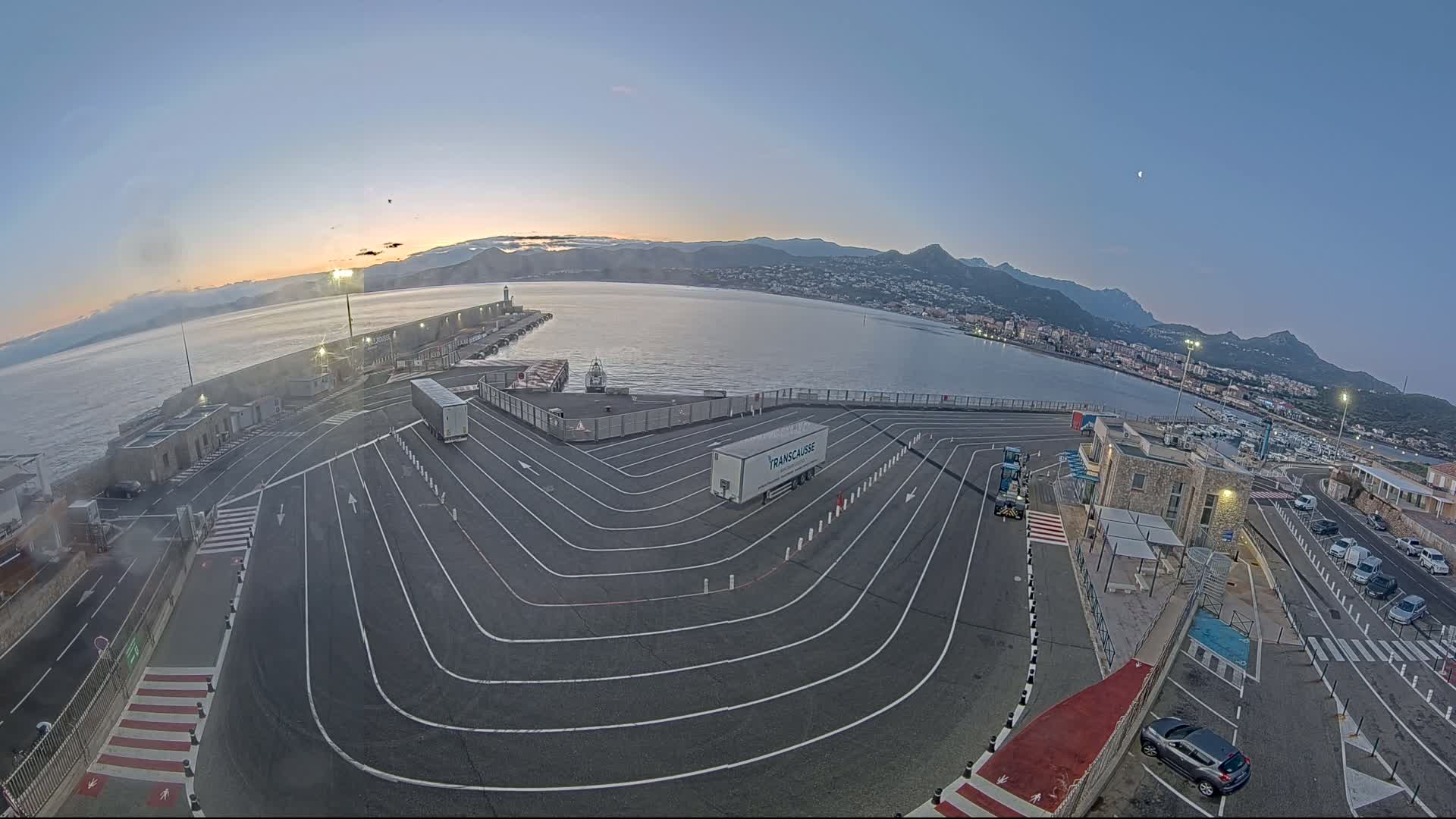 A ferry terminal with a large, curved roadway marked with white lines, a few vehicles, and a calm body of water with mountains in the background under a twilight sky.