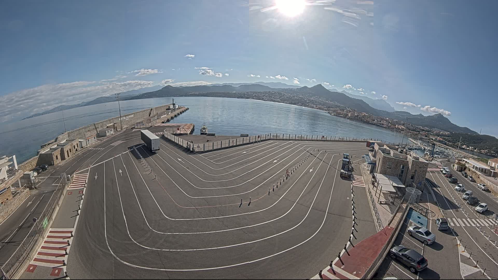 A sunny day at a harbor shows a large, empty paved area with marked lanes curving toward a dock, a body of water with mountains in the background, and several cars parked nearby.