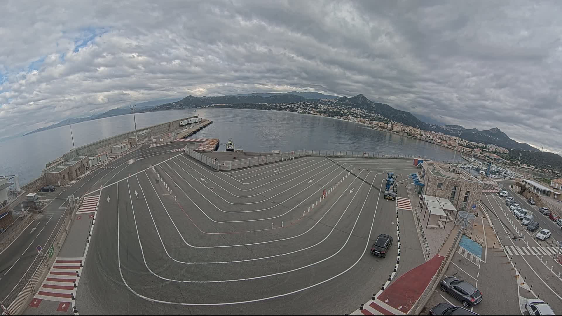 An aerial view of a ferry terminal with a large, marked queuing area, overlooking a calm bay under a cloudy sky.