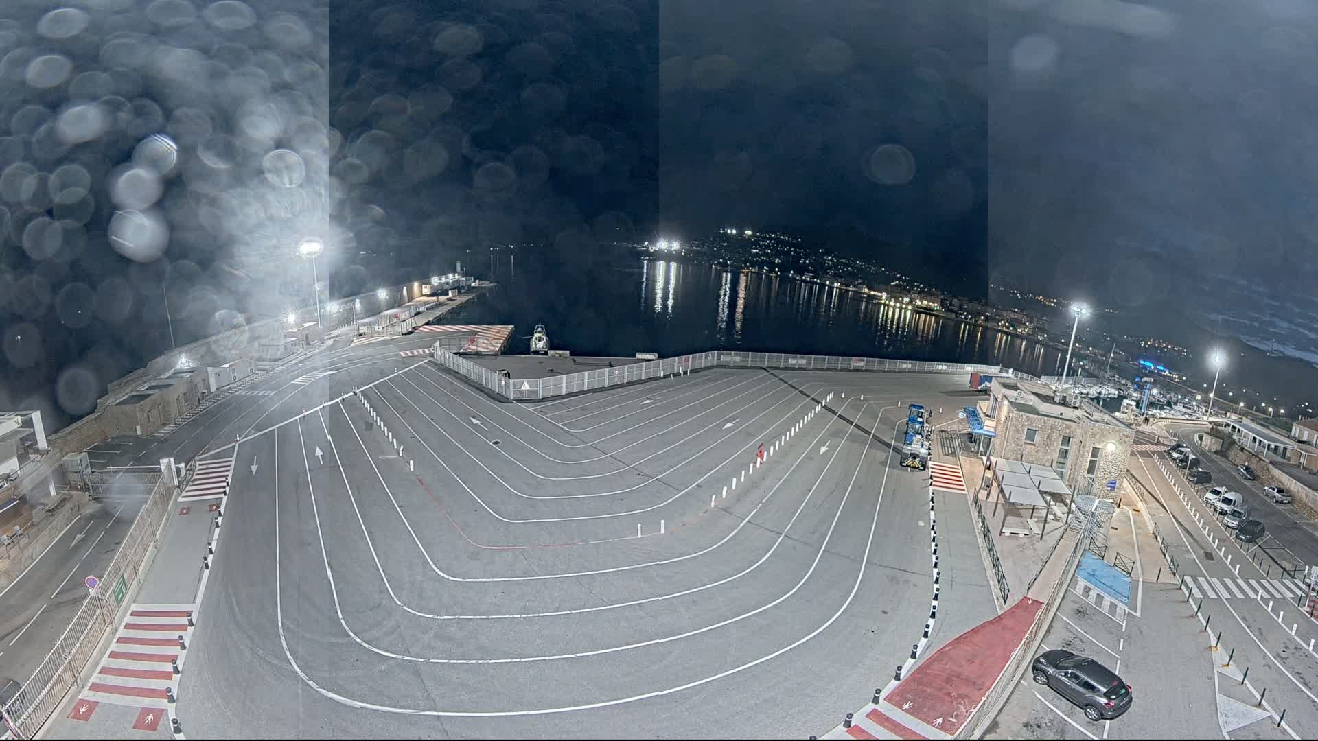 An empty ferry terminal at night, under a dark, slightly overcast sky, shows a large, marked area for vehicles next to a calm body of water with lights visible across the bay.