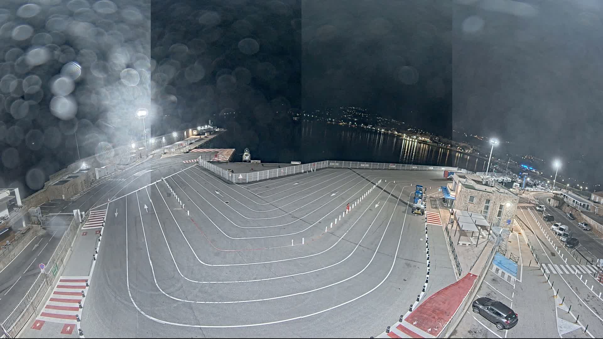 A nighttime view of an empty ferry terminal with marked lanes and a calm body of water in the background, seen through rain-streaked glass.