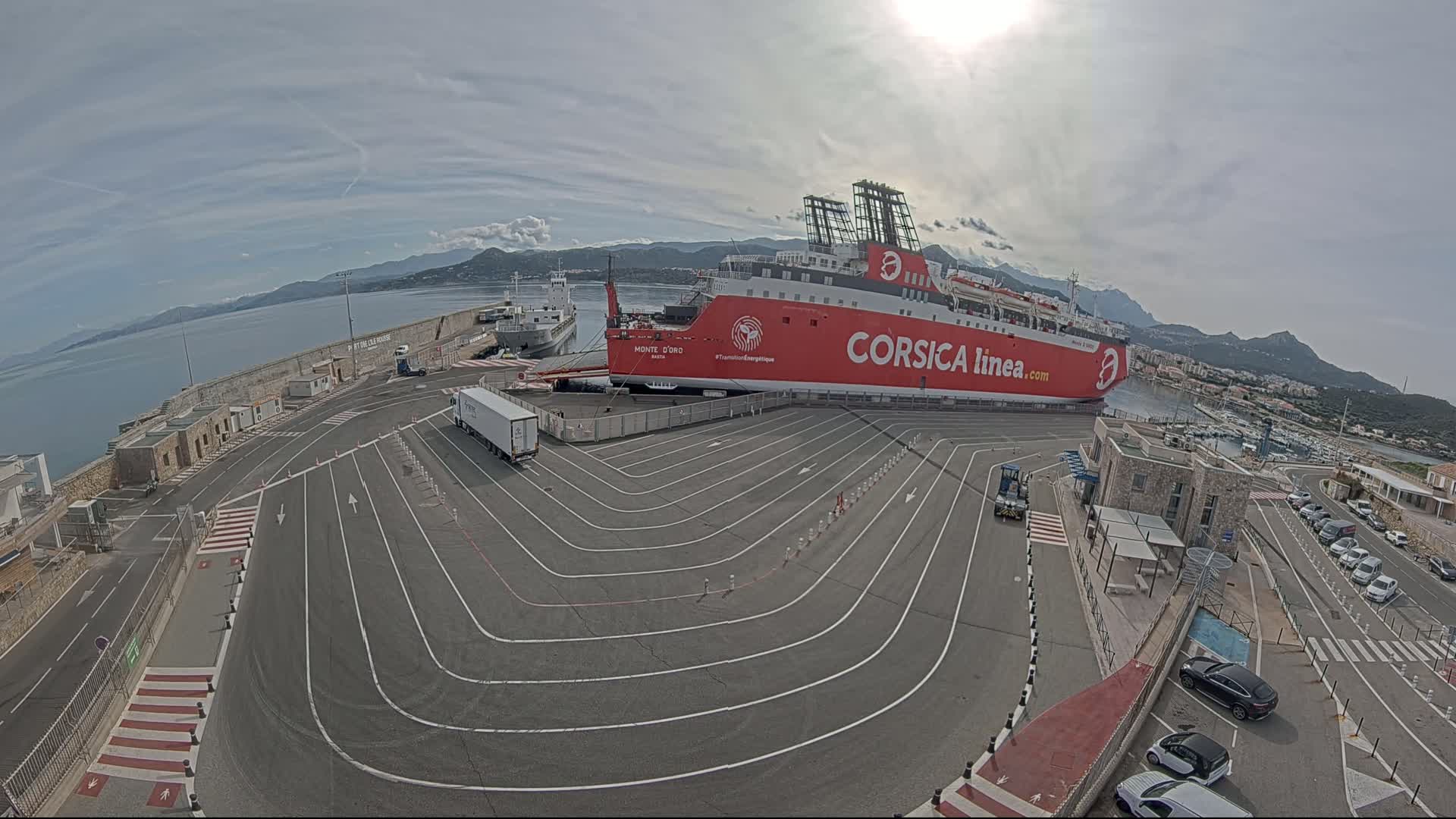 A large red ferry is docked at a port on a sunny day, with several cars parked nearby and a truck on the dock.