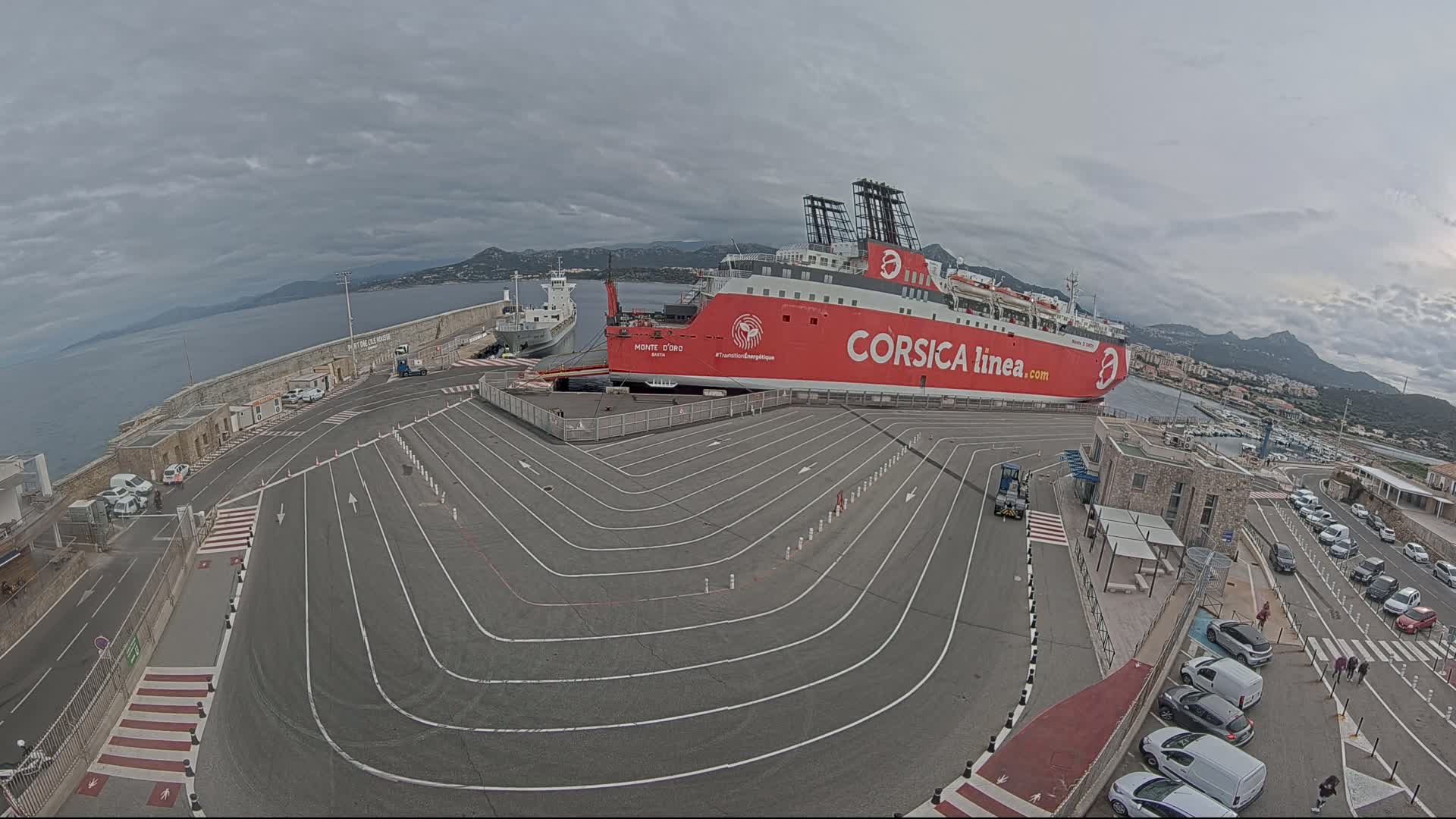 A large red ferry is docked at a port on a cloudy day, with a smaller cargo ship nearby and several cars parked in the surrounding area.