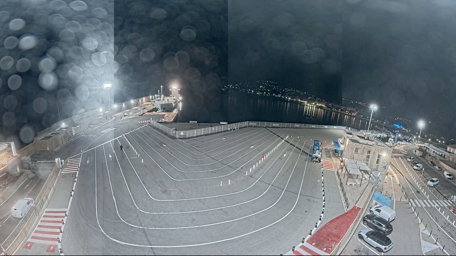 A nighttime view of a ferry terminal with a ship docked, a large empty vehicle queuing area with painted lane markings, and several cars parked nearby, all seen through rain-covered glass.
