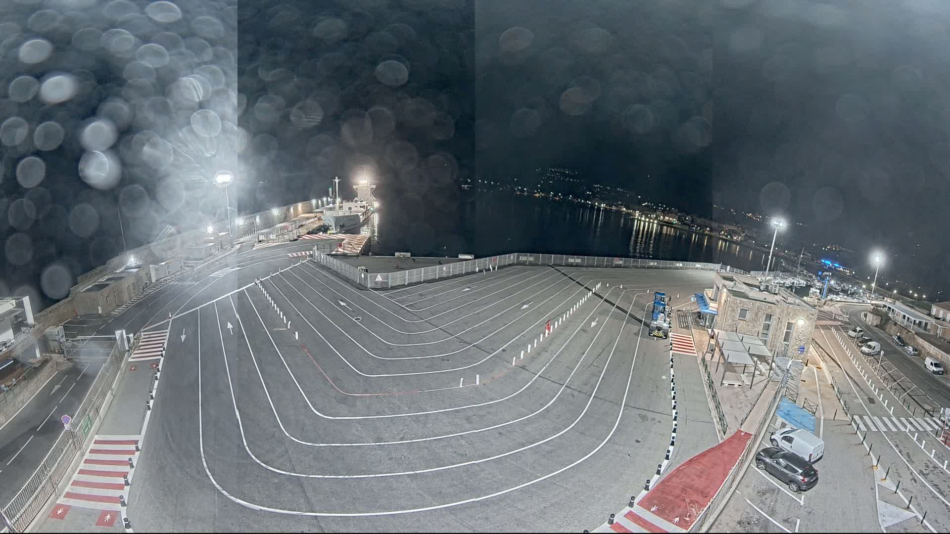 A nighttime, rain-streaked view of a ferry terminal with a large, lined parking area, a docked ship, and a small building.