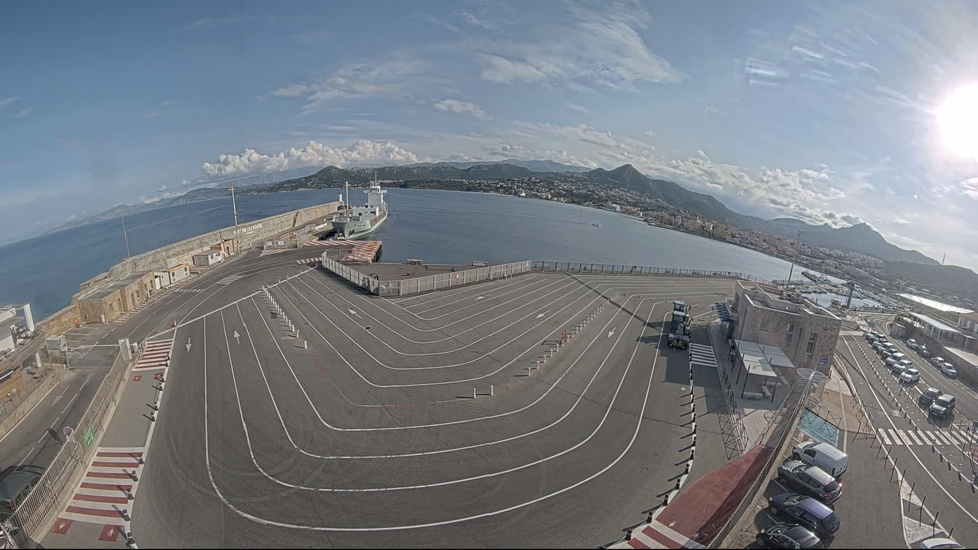 A large, empty ferry terminal with marked lanes curves toward a docked ship and body of water under a mostly sunny sky, mountains in the distance.