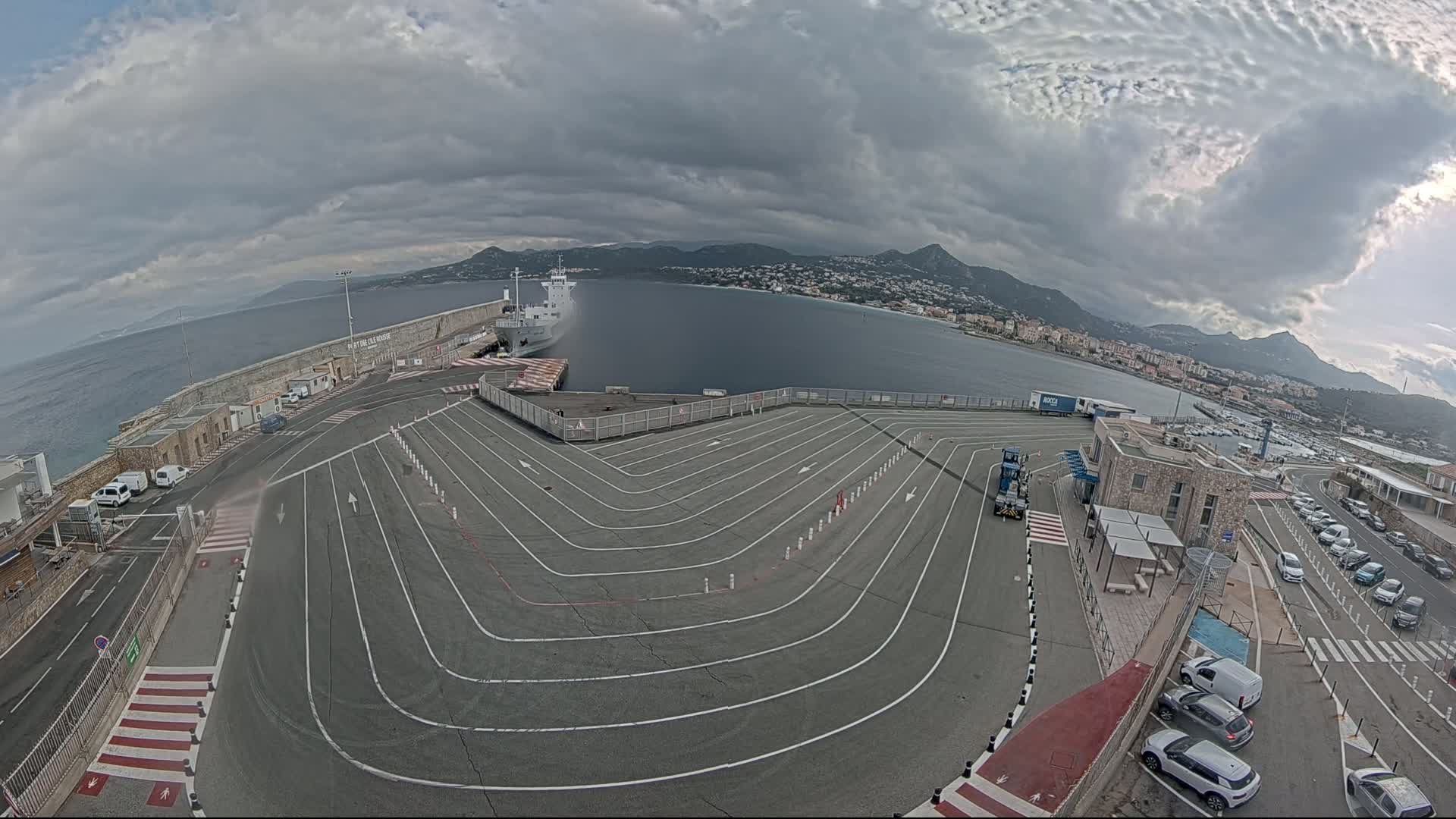 A large, empty ferry terminal with marked lanes curves toward a docked ship and calm ocean water under a cloudy sky.