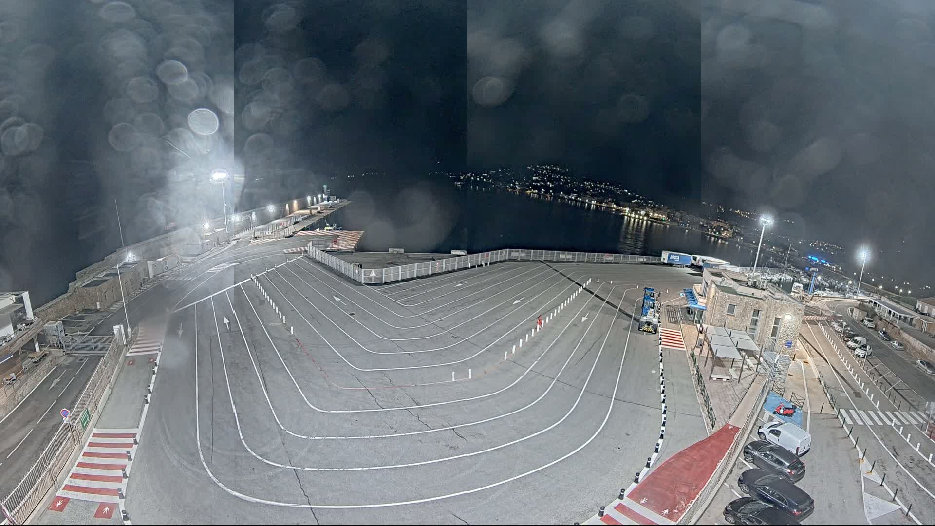 A nighttime, rain-streaked view of a ferry terminal's empty vehicle lanes curving toward a waterfront, with a few cars parked nearby and a town's lights visible across the water.