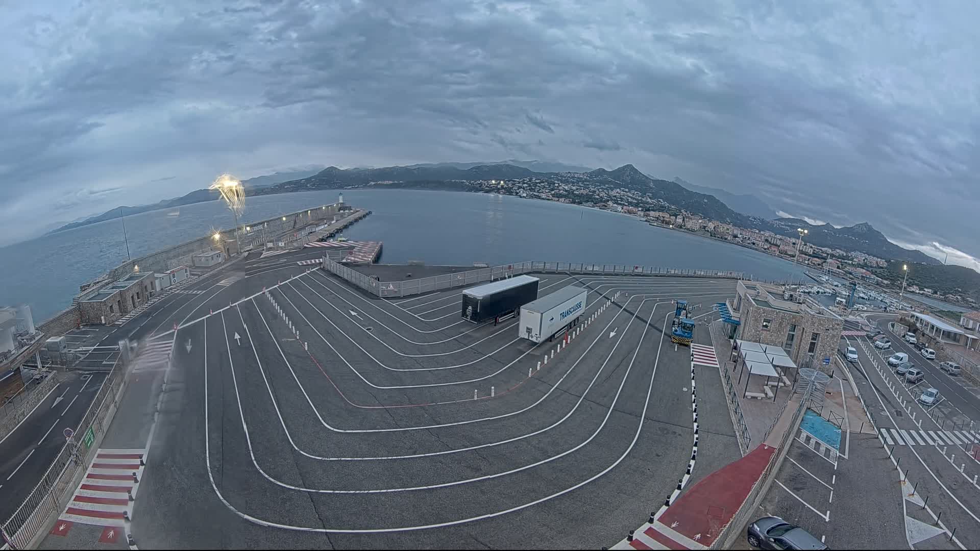 An aerial view of a port on a cloudy day shows a large empty parking lot with lane markings, two semi-trucks, and a few cars parked near buildings, all overlooking a calm body of water and distant mountains.