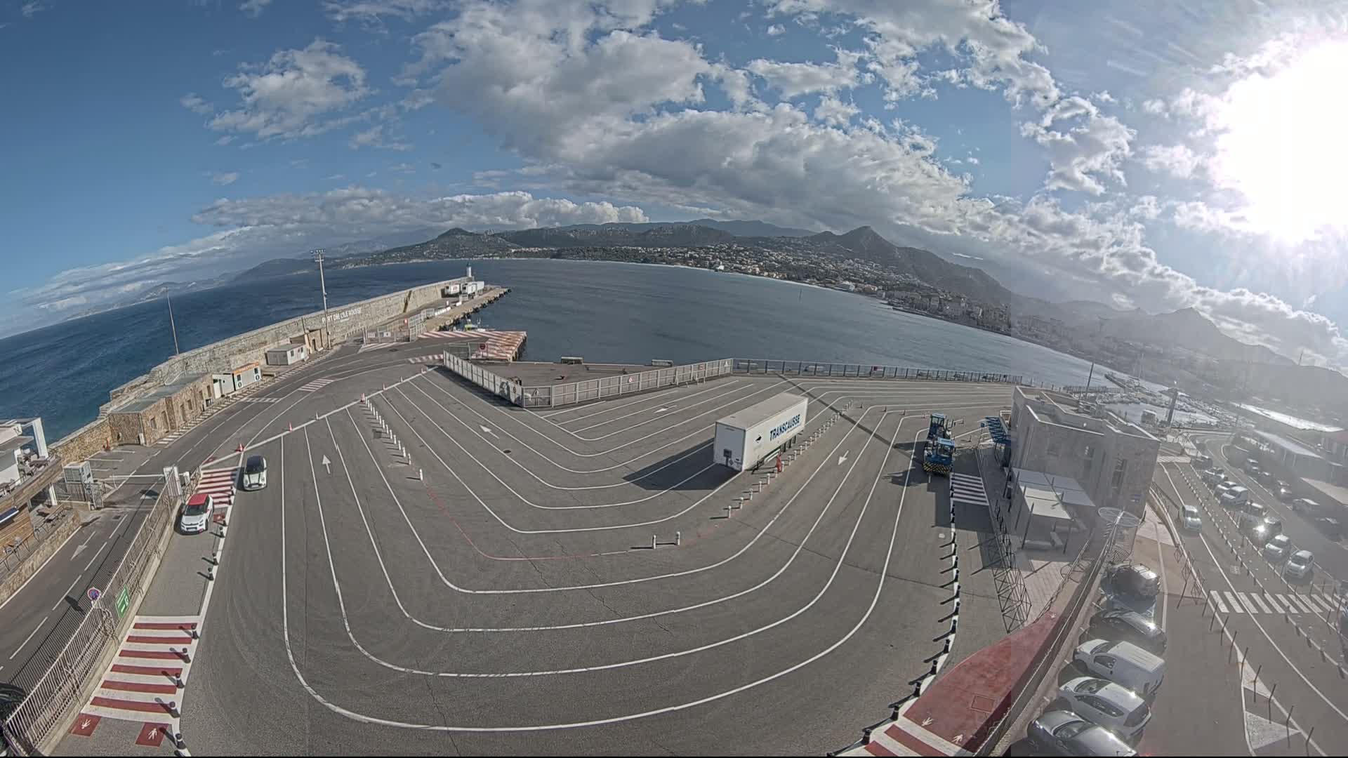 A mostly sunny day overlooks a port with a large, curving vehicle staging area, a body of water, and mountains in the background.