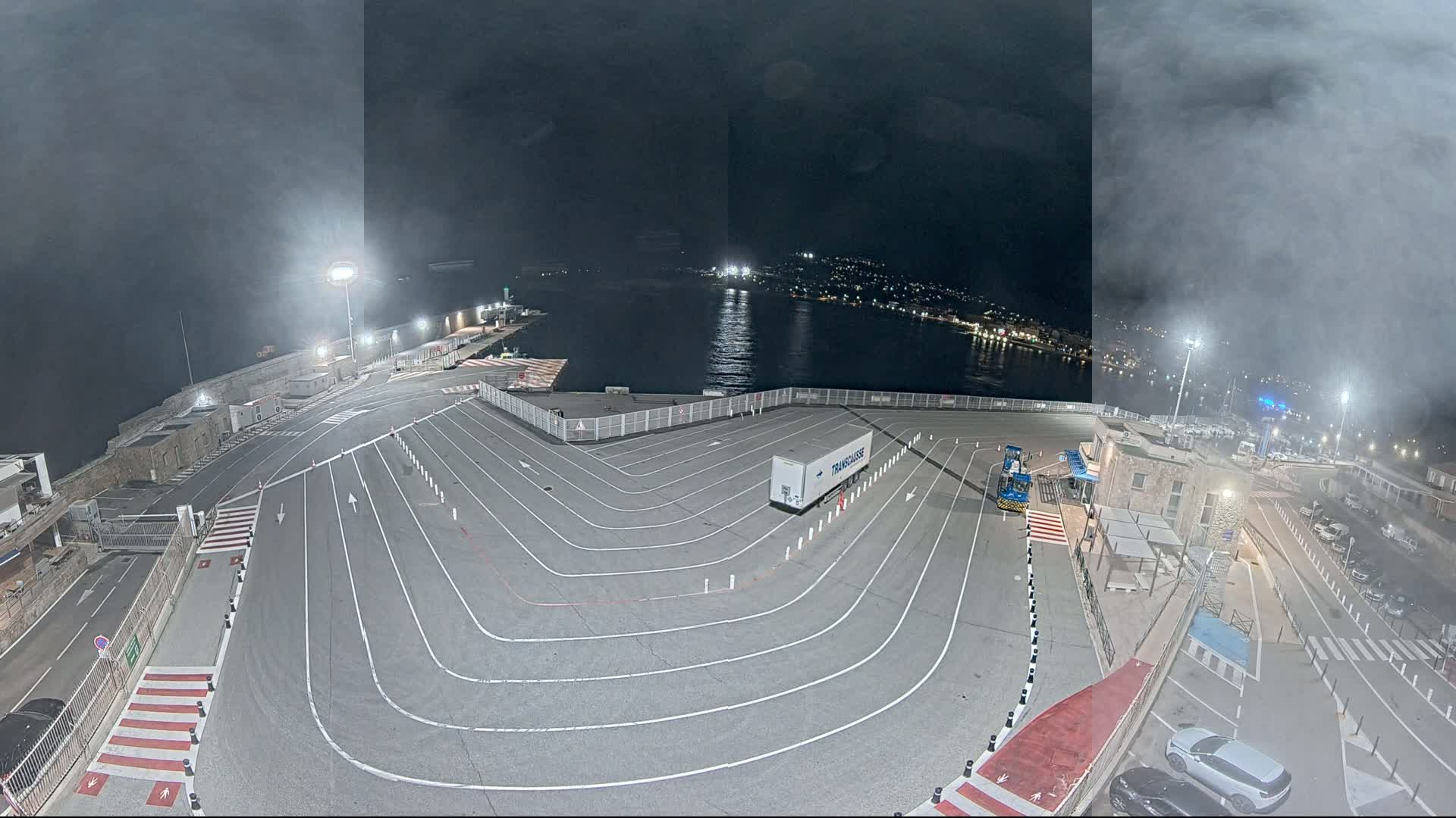 A nighttime, hazy view of a ferry terminal with a large, marked vehicle staging area and a single semi-trailer truck parked near a body of water.