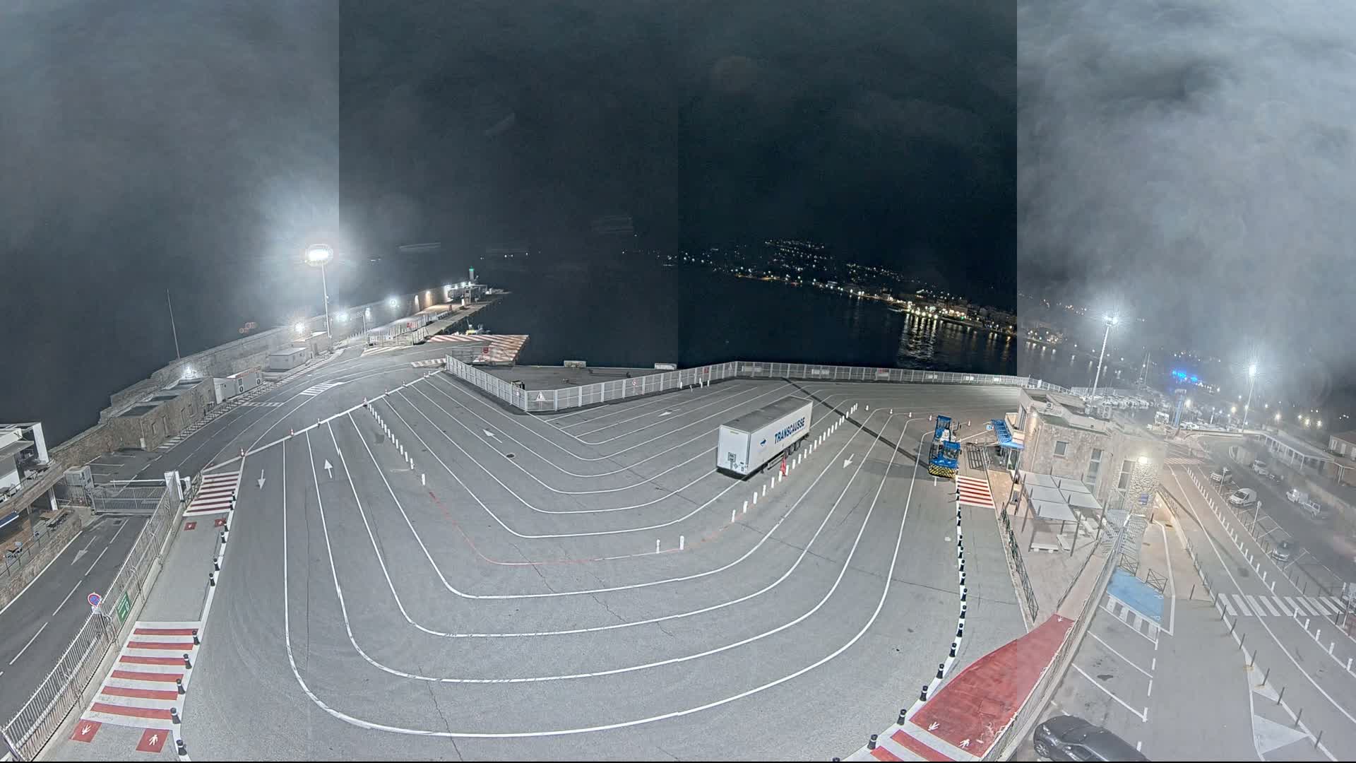 A nighttime, foggy view of a port's vehicle staging area with a single large trailer parked within marked lanes, overlooking a dark body of water with city lights in the distance.