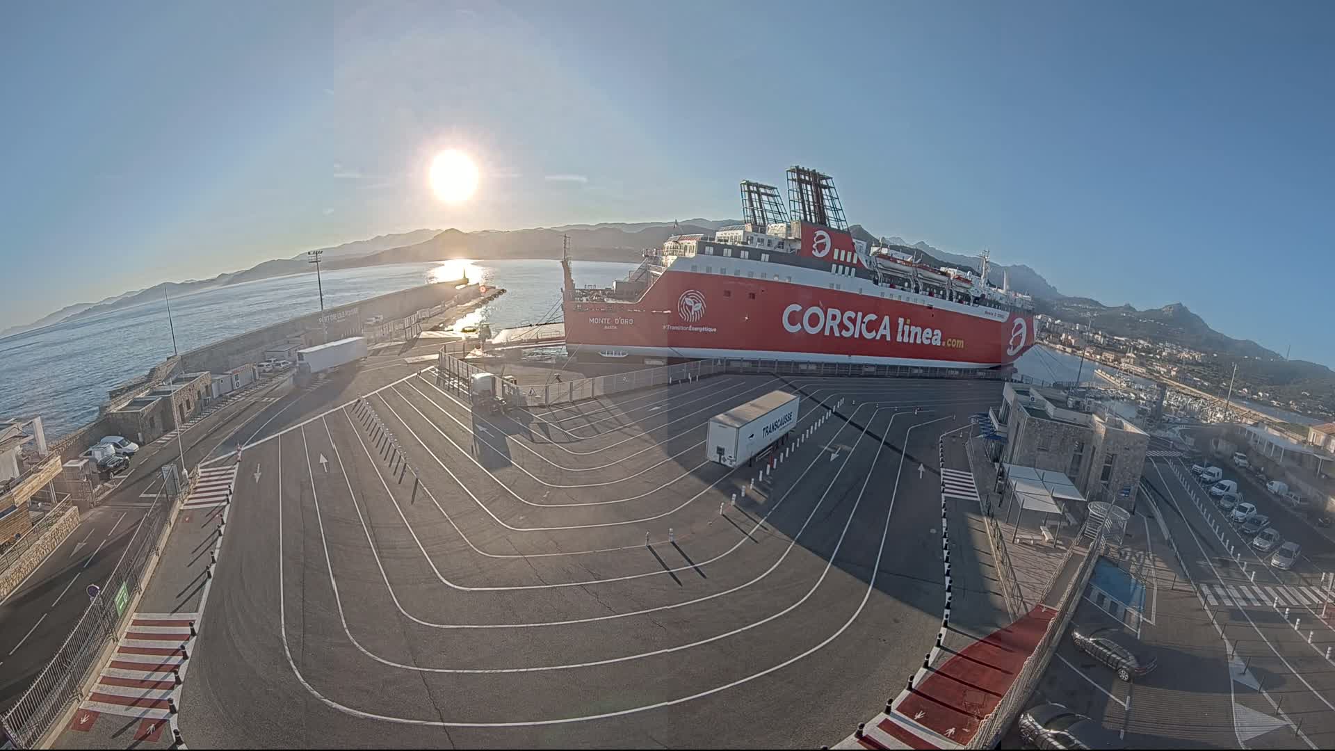 A large red ferry is docked at a port on a sunny day, with a calm body of water and mountains in the background.