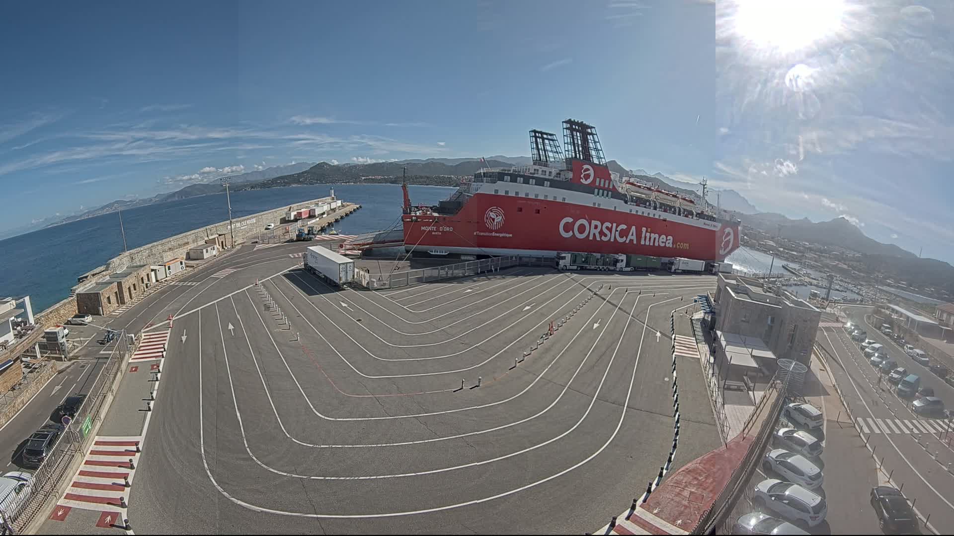 A large red ferry is docked at a port on a sunny day, with a paved area marked with driving lanes leading to it.