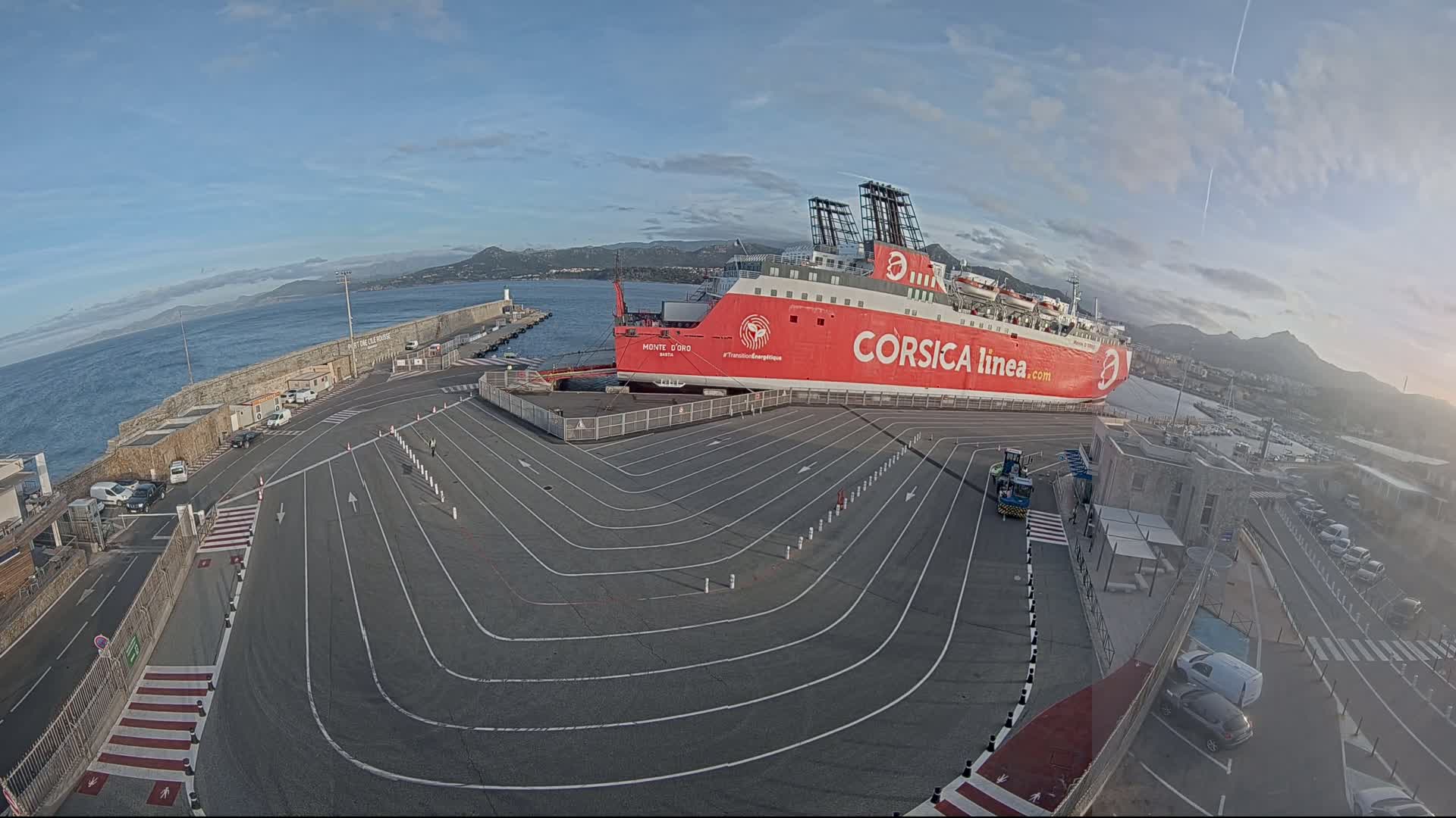 A large red ferry is docked at a port under a mostly clear sky, with a paved area marked with driving lanes surrounding it.