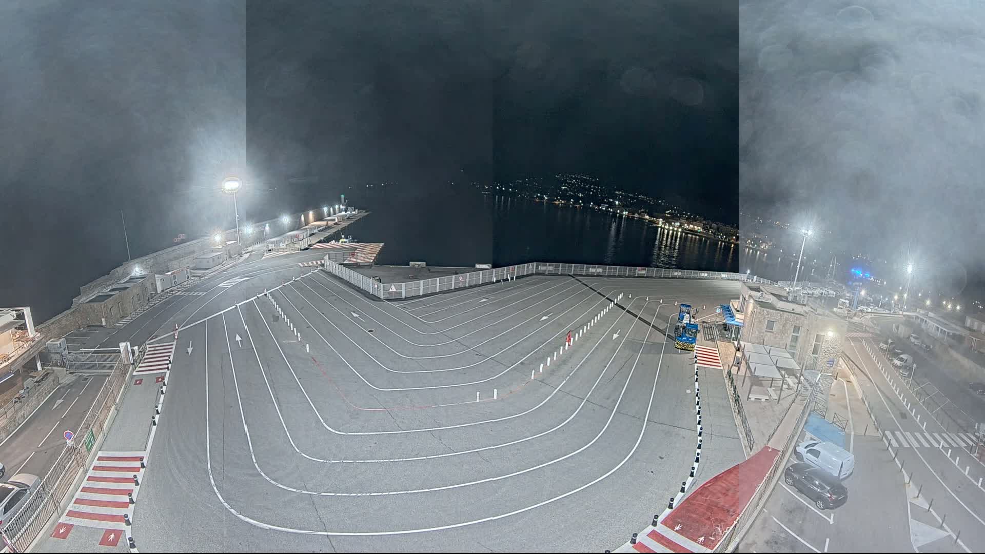 An empty, marked vehicle staging area at a nighttime harbor is shrouded in fog or mist, overlooking calm water and a city's lights.