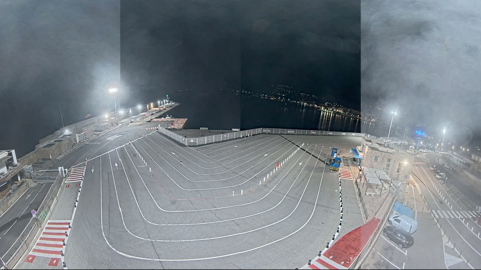 A nighttime, hazy view of a large, empty vehicle staging area with marked lanes next to a body of water.