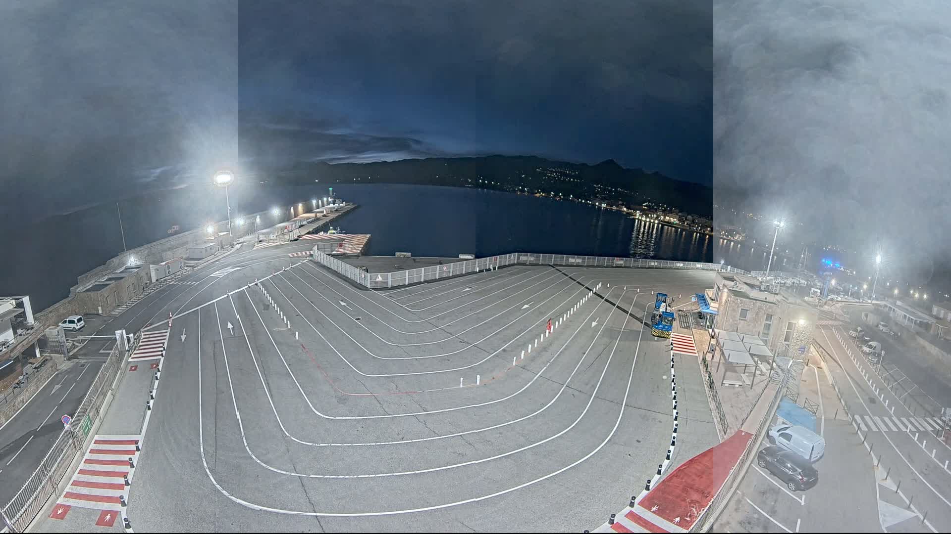 An empty, marked vehicle staging area at a harbor at night, under a hazy sky.